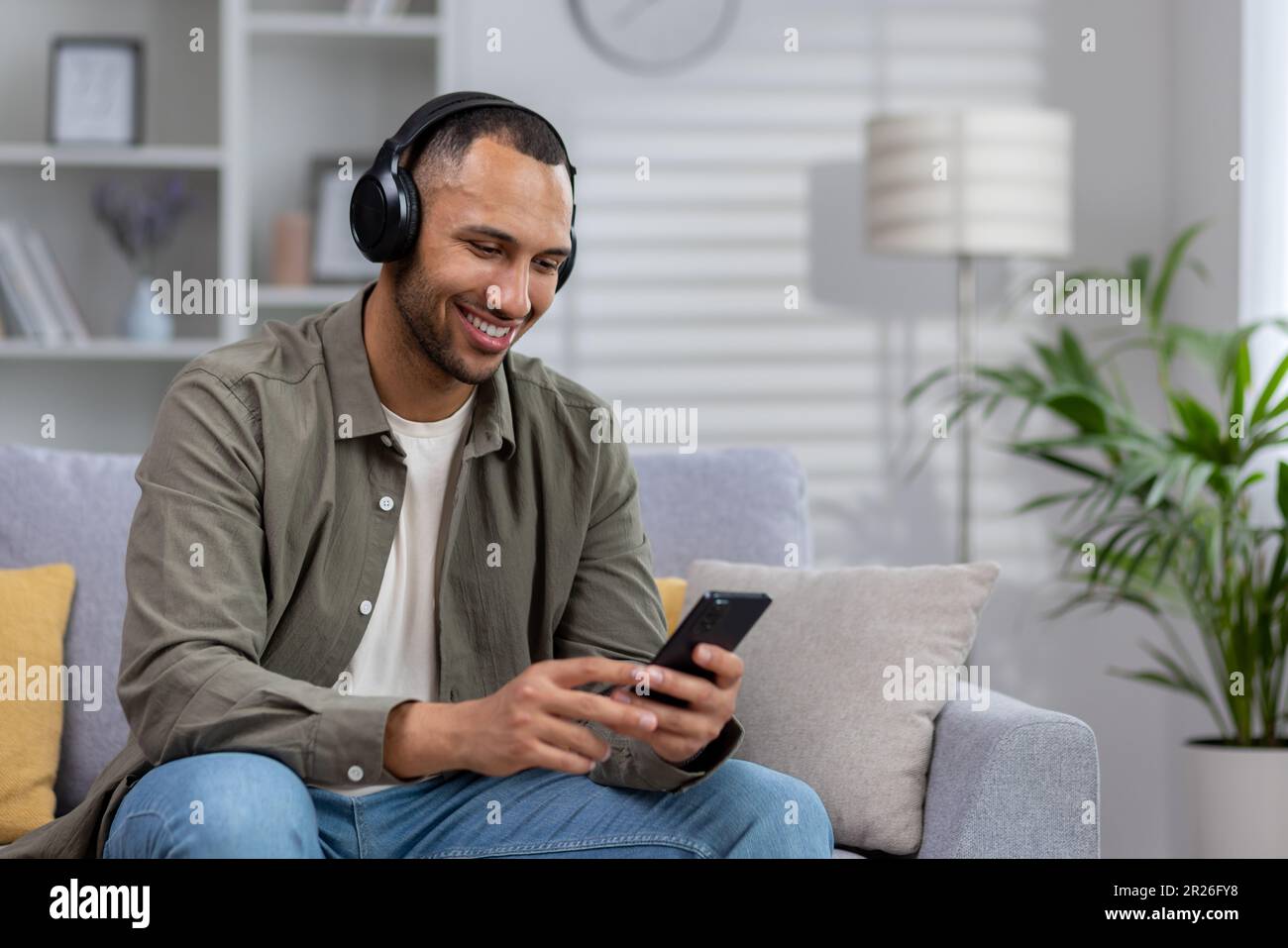 Young hispanic man at home sitting on sofa resting, man with headphones listening online music, radio and audio books podcasts in app on phone. Stock Photo