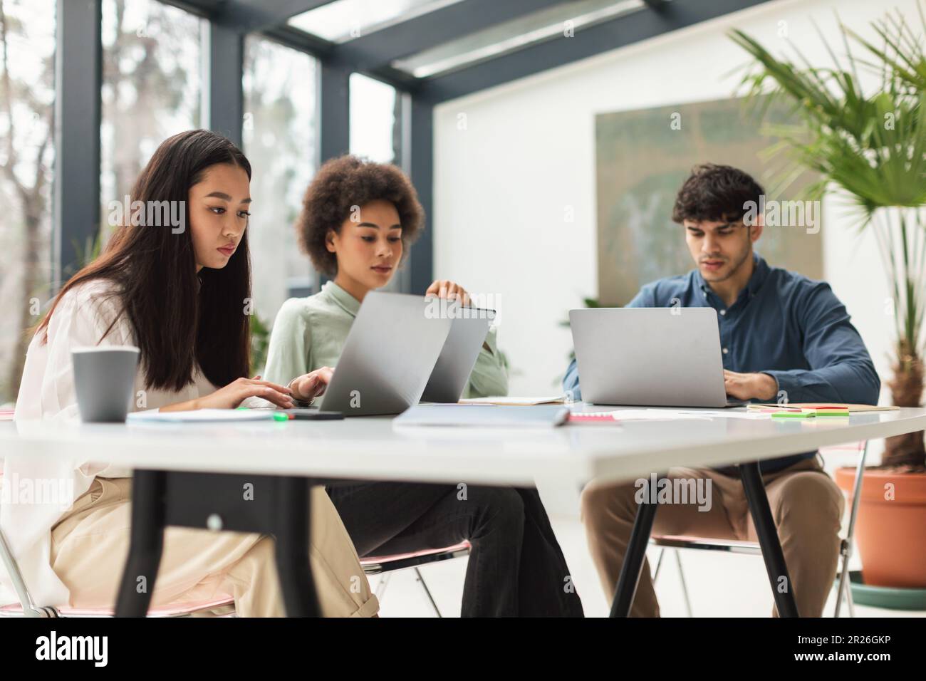 Three Multiethnic Coworkers Working Using Laptops At Table In Office Stock Photo