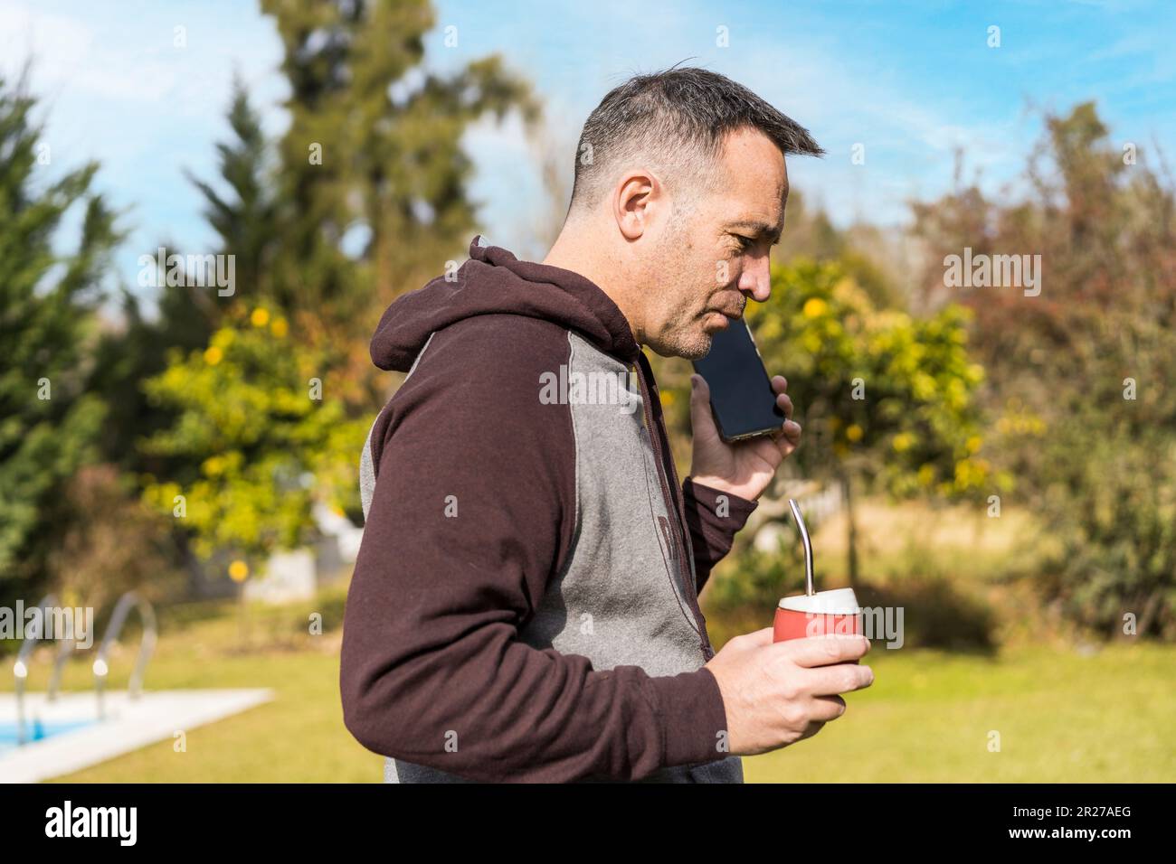 Adult man listening to messages on his phone and drinking mate infusion while walking in his garden.  Home office concept. Stock Photo