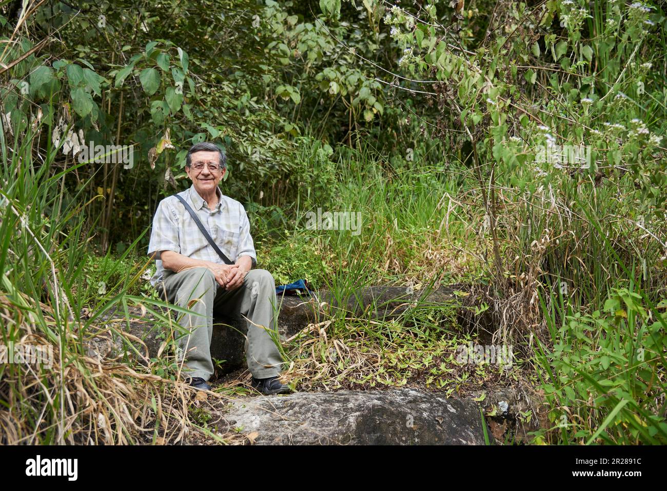 Male senior traveler sitting, smiling and looking at the camera, taking a break during a hike in a rural setting near Aratoca in Santander, Colombia. Stock Photo