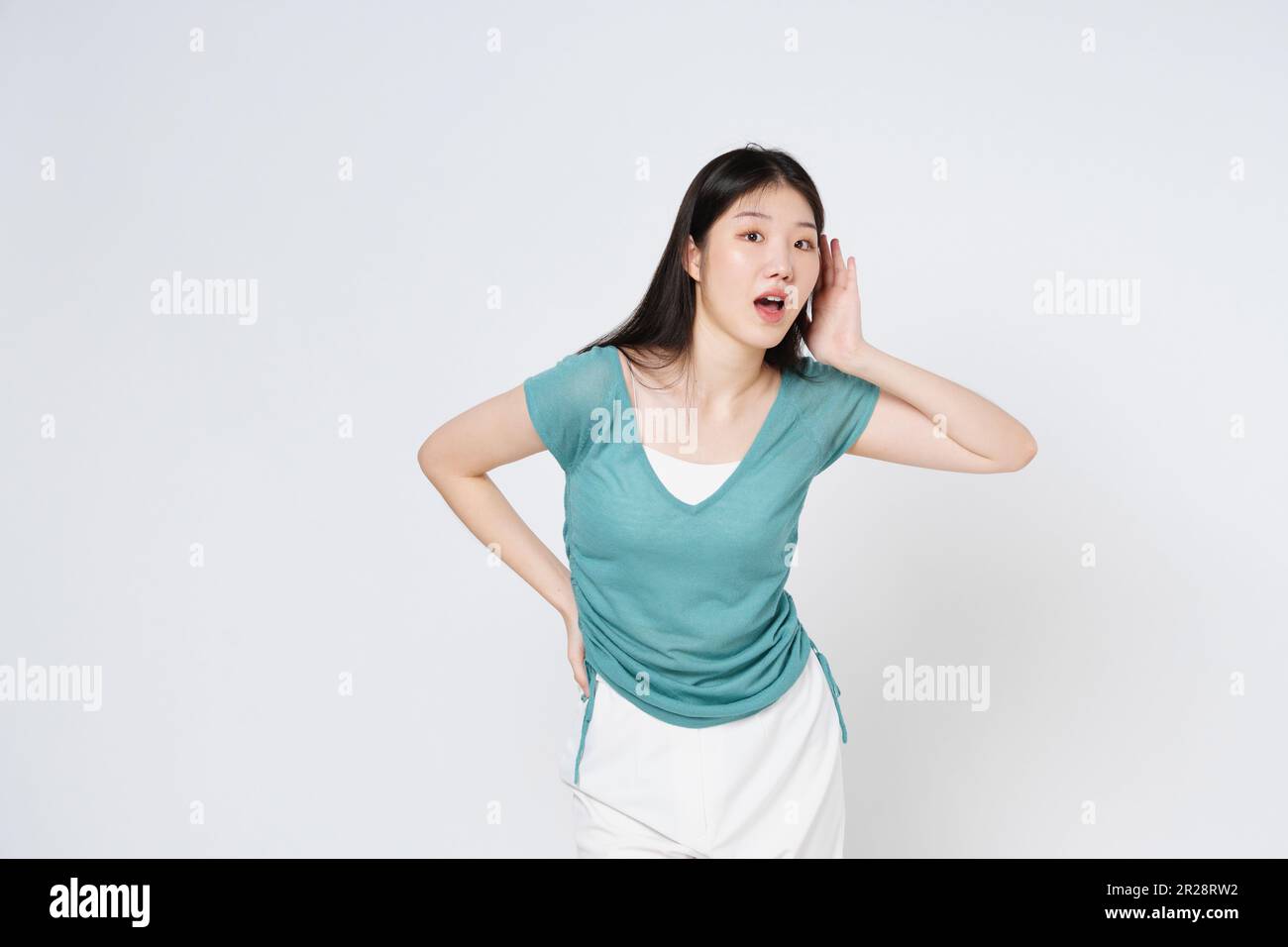 Young woman with opened mouth trying to hear something with raised hand near her ear and looking at camera, while standing over white isolated backgro Stock Photo