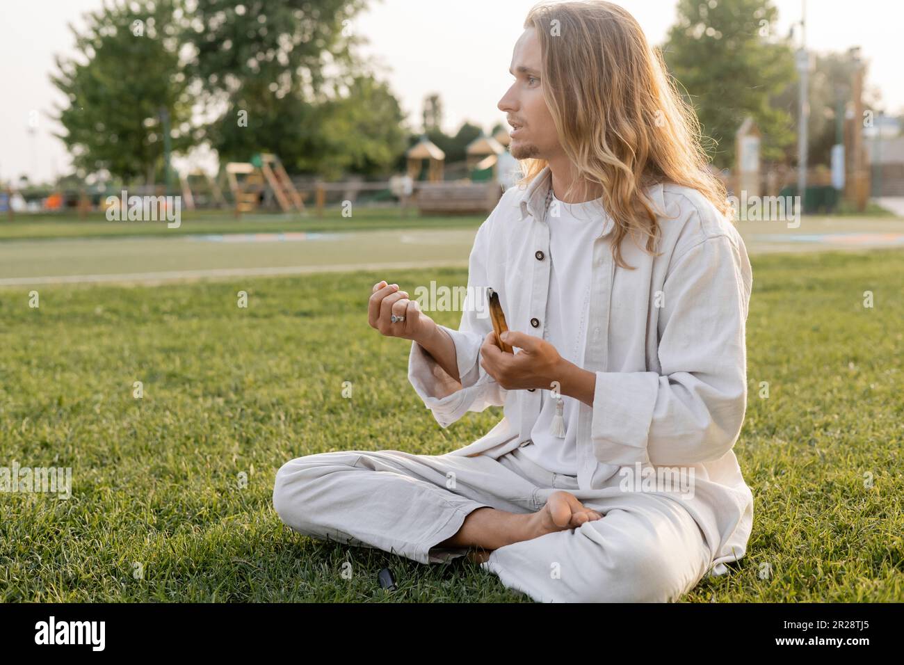 barefoot man in white linen clothes meditating in easy pose with smoldering palo santo stick and looking away outdoors Stock Photo