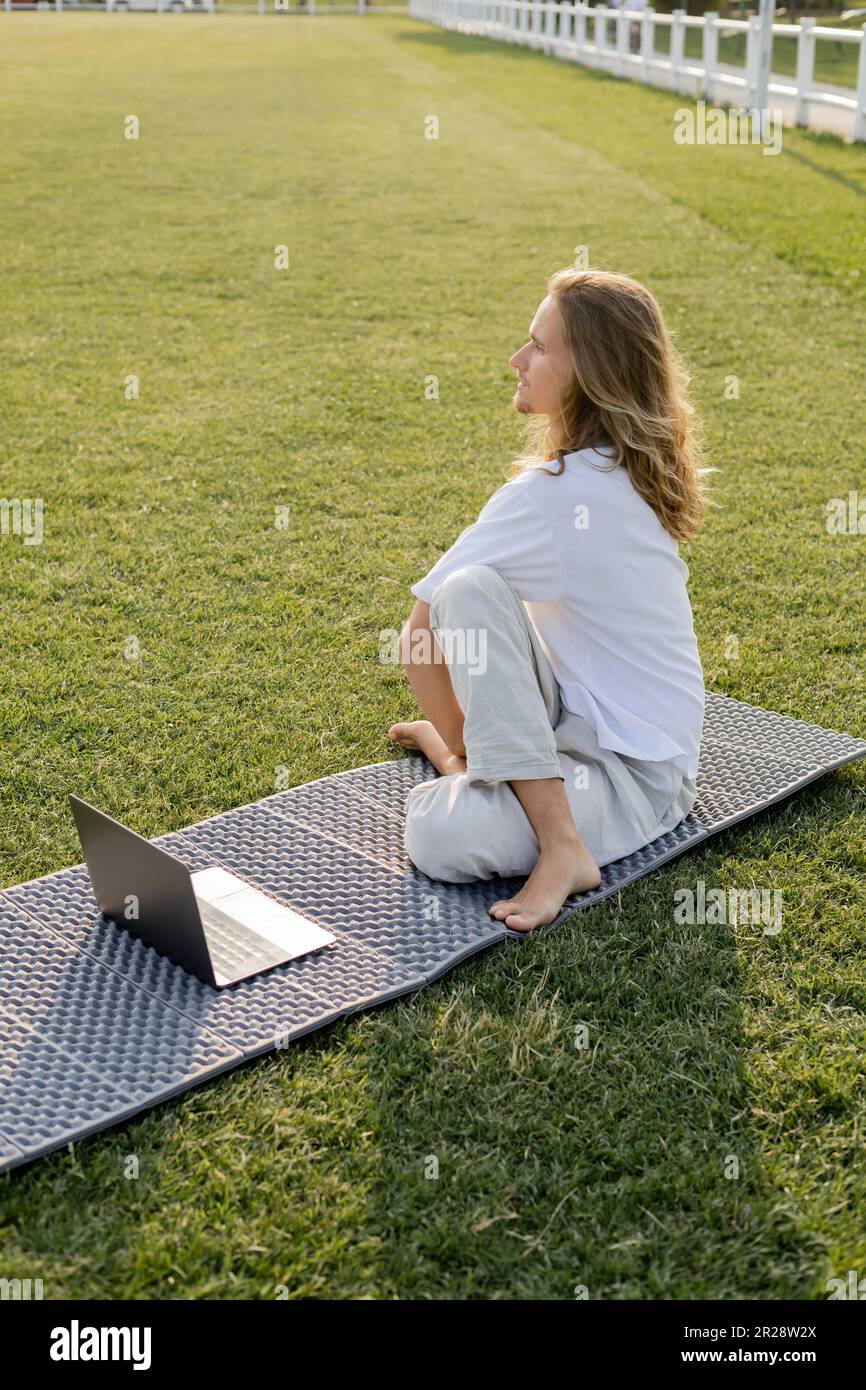 long haired man sitting in sage pose near laptop and looking away on yoga mat on green field Stock Photo