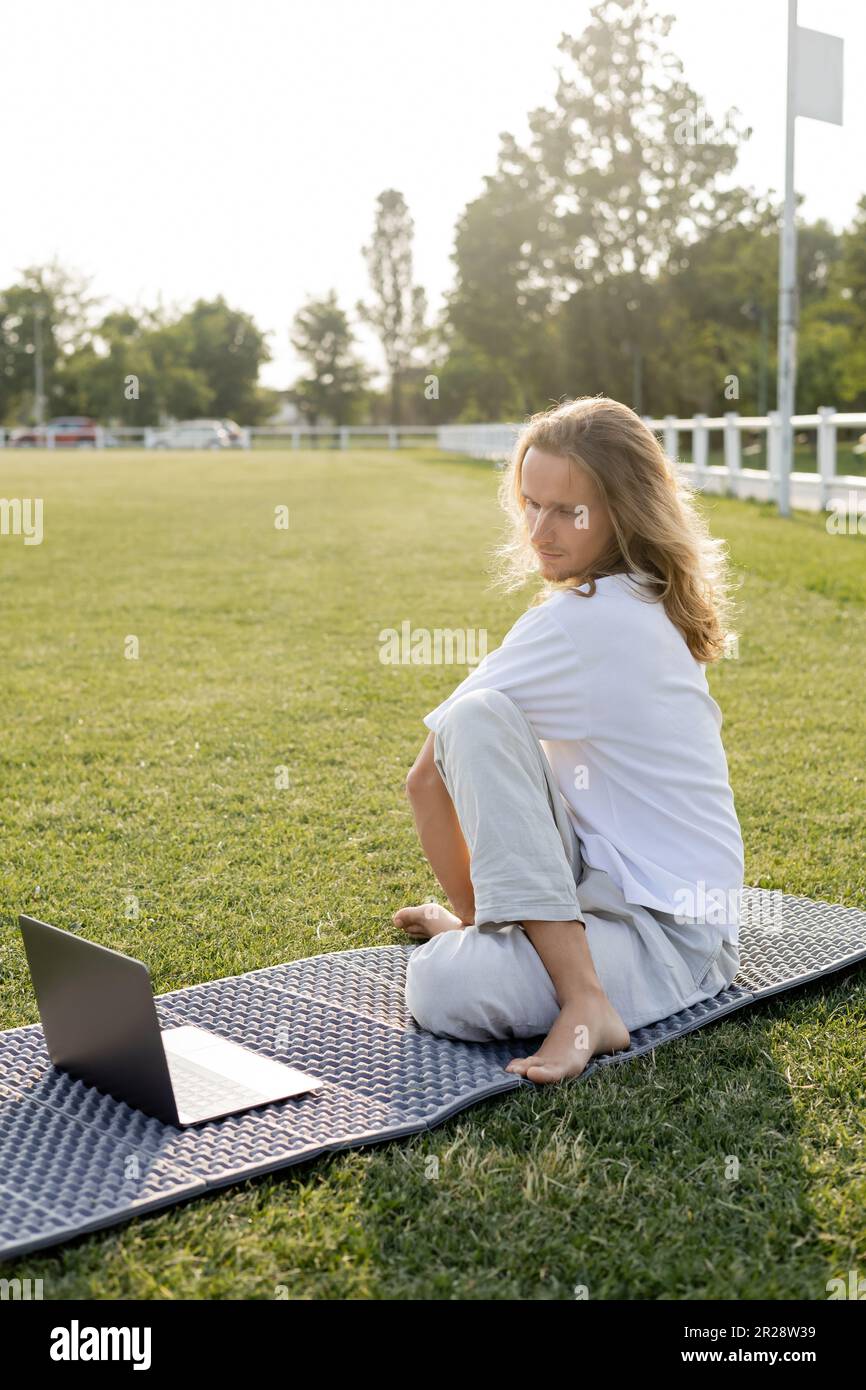 long haired man sitting in sage pose during online lesson on laptop on green lawn of outdoor stadium Stock Photo