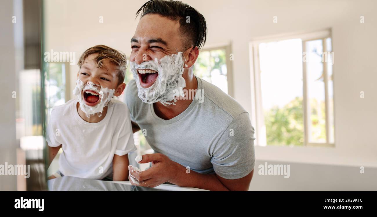 Father and son having fun in a bathroom, laughing happily with shaving foam on their faces. Young single dad taking a moment to bond and share moments Stock Photo