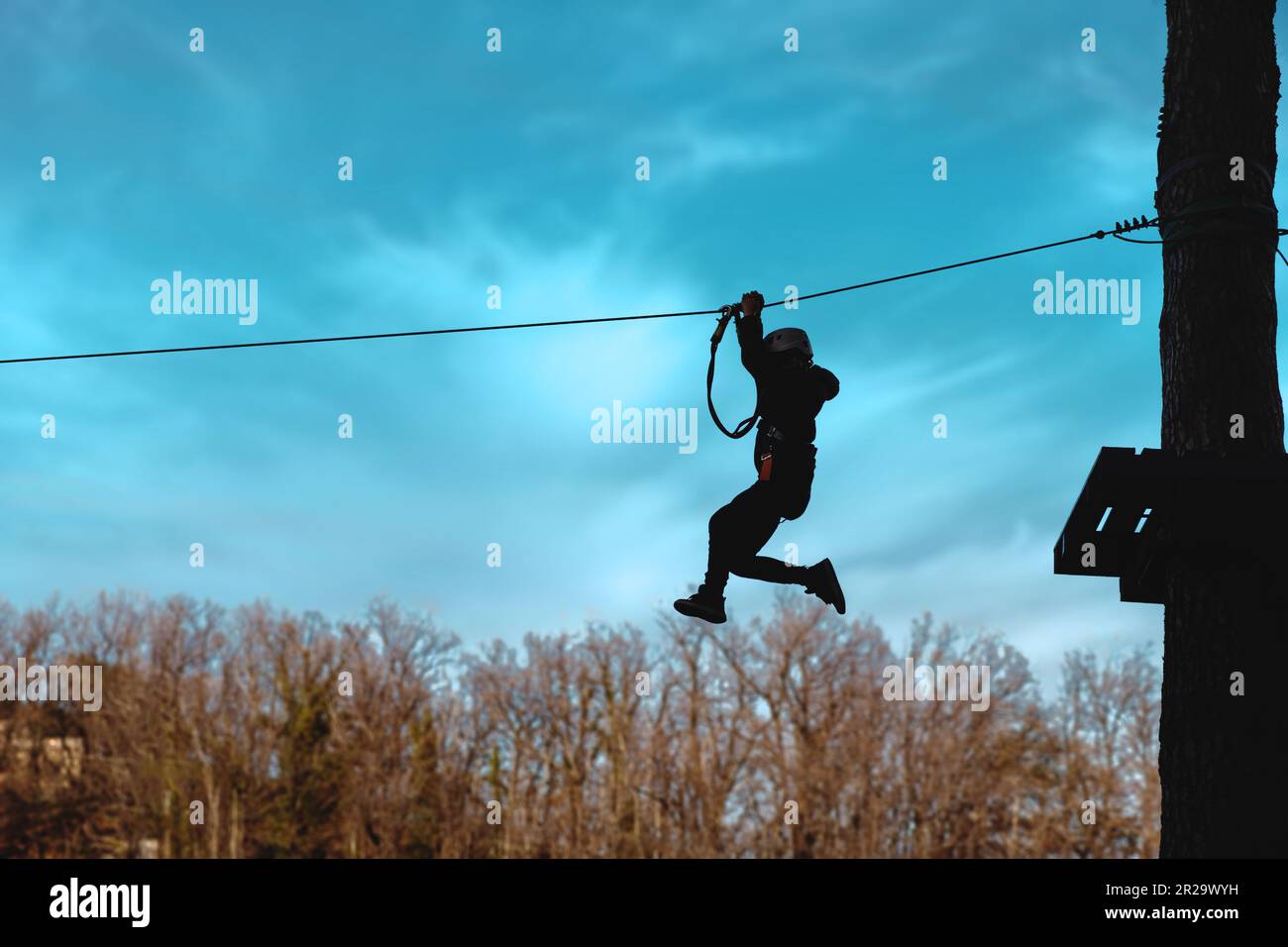 Silhouette of an unrecognizable person zip-lining at an adventure park. Wearing a safety helmet and harness, they are captured mid-air against a blue Stock Photo