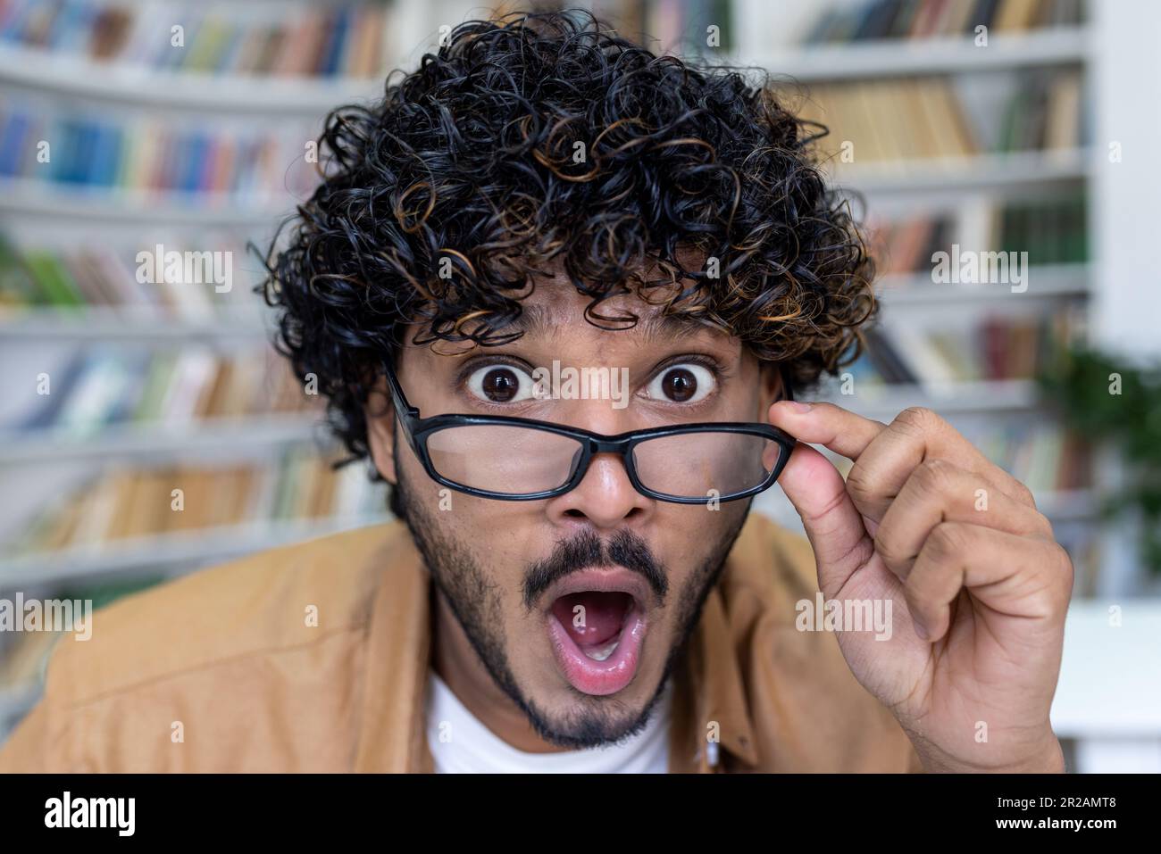 Close-up photo. Portrait of a young hispanic man in a library, bookstore, who looks at the camera in surprise, lowers his glasses on his face. Stock Photo
