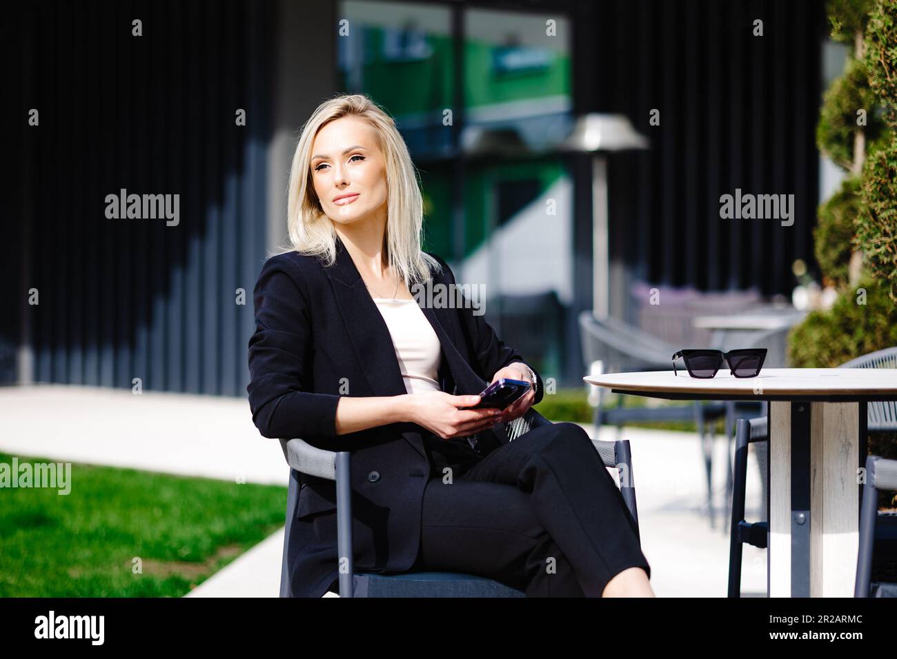 Blonde business lady looks at the phone, reading messages. Woman is relaxing in a cafe on the terrace enjoyment of life. Stock Photo