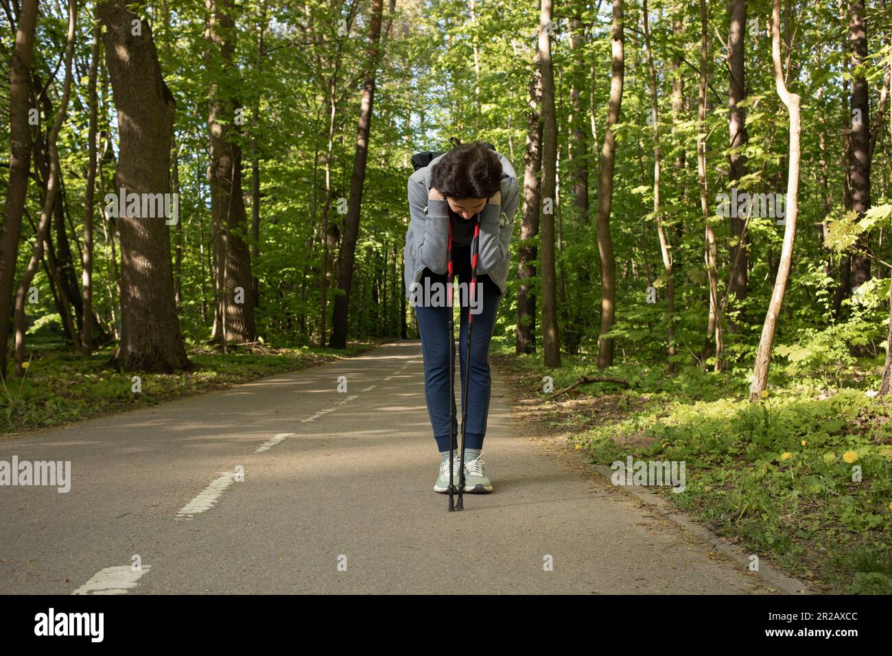 Woman bends over leaning walking poles feeling pain, not well while doing sport, jogging, Nordic walk in Park.Hard breathing, Dizziness. Sickness Stock Photo