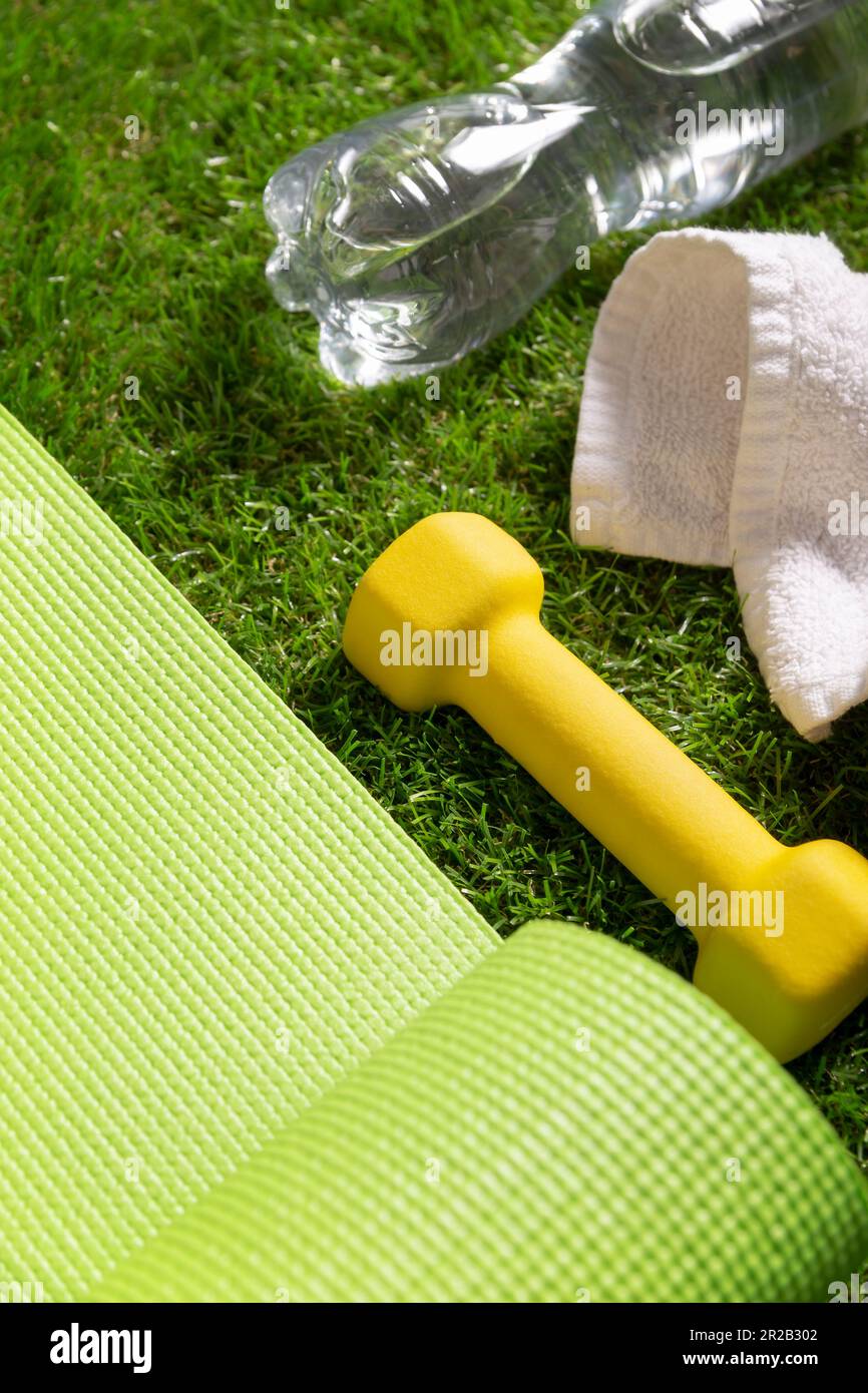 Fitness, hydration, exercise, healthy and active lifestyles concept image. Rolled up green yoga mat, yellow dumbbell and water bottle on green grass i Stock Photo