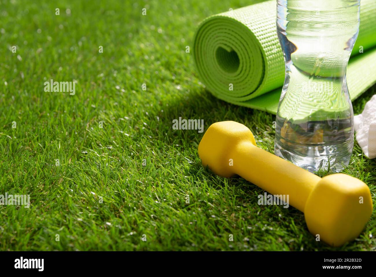 Fitness, hydration, exercise, healthy and active lifestyles concept image. Rolled up green yoga mat, yellow dumbbell and water bottle on green grass i Stock Photo