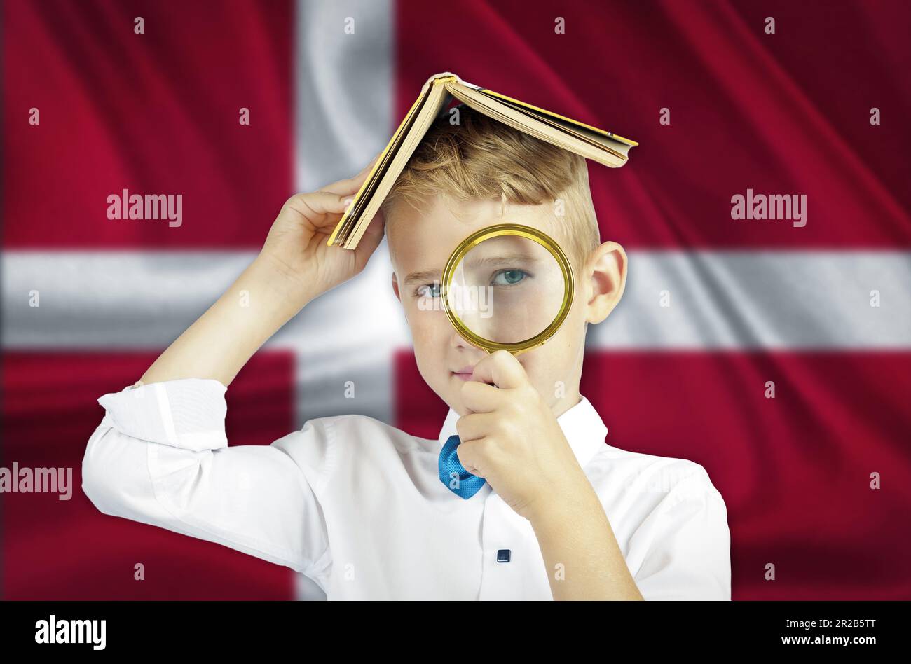 A boy with a book on his head looks through a magnifying glass at the background of the Danish flag.Education concept. Stock Photo