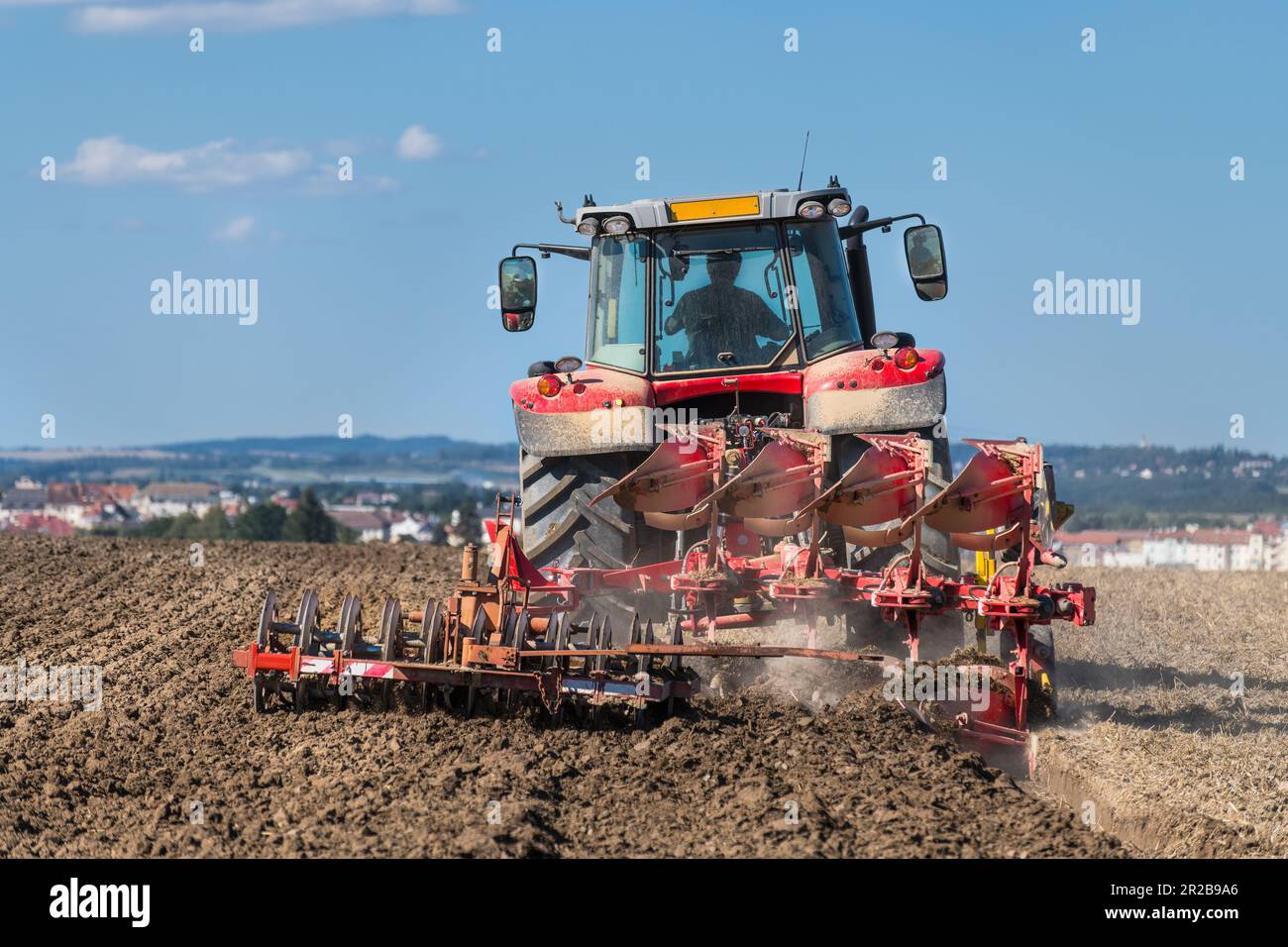 Red tractor on field at plowing and harrowing farm land soil in summer nature under blue sky. Working farmer in agricultural vehicle at earth tillage. Stock Photo
