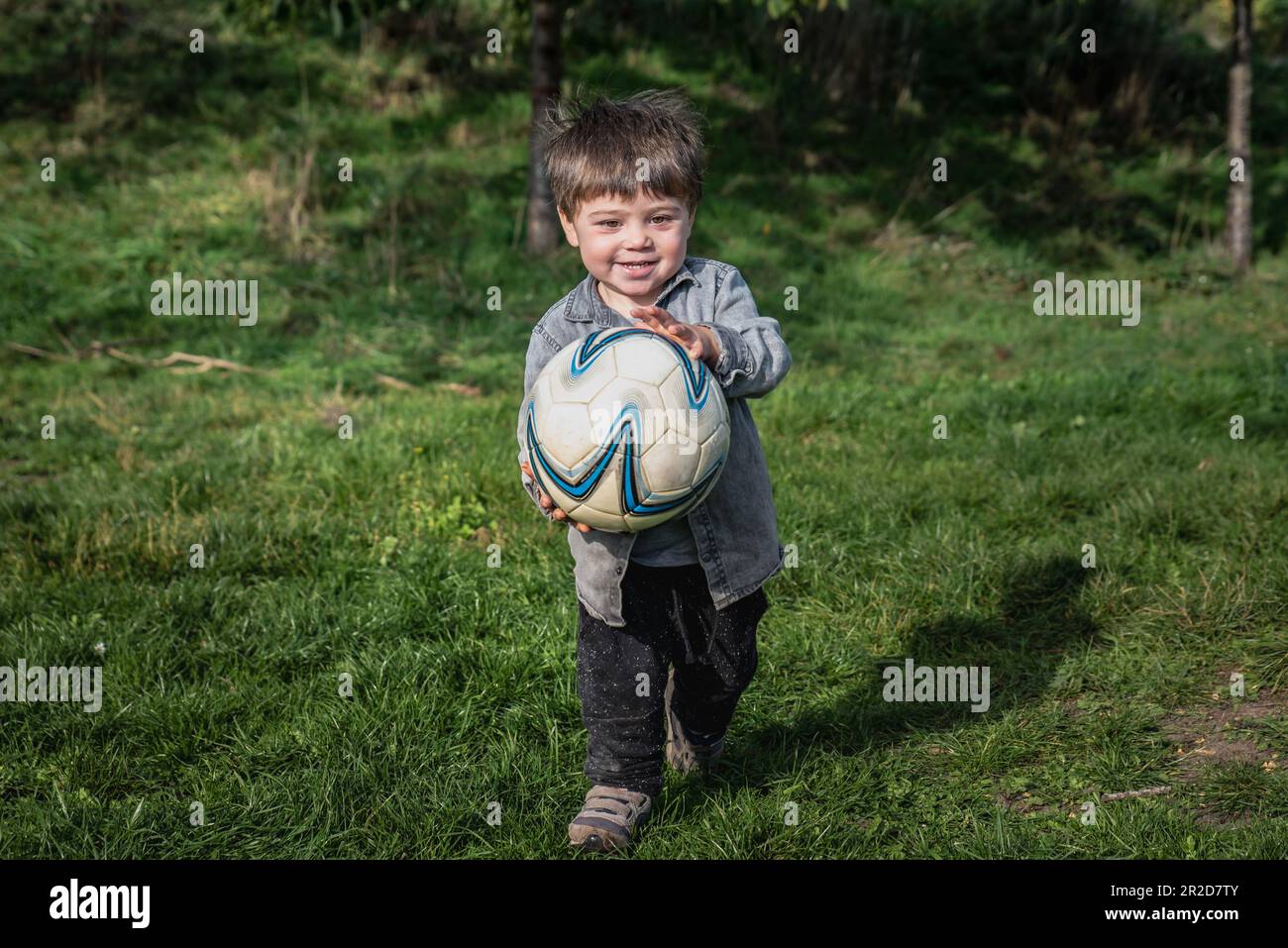 Happy kid carrying a big soccer ball is ready to play. Smiling boy engaging in an outdoor football activity conveys a healthy growth and lifestyle Stock Photo