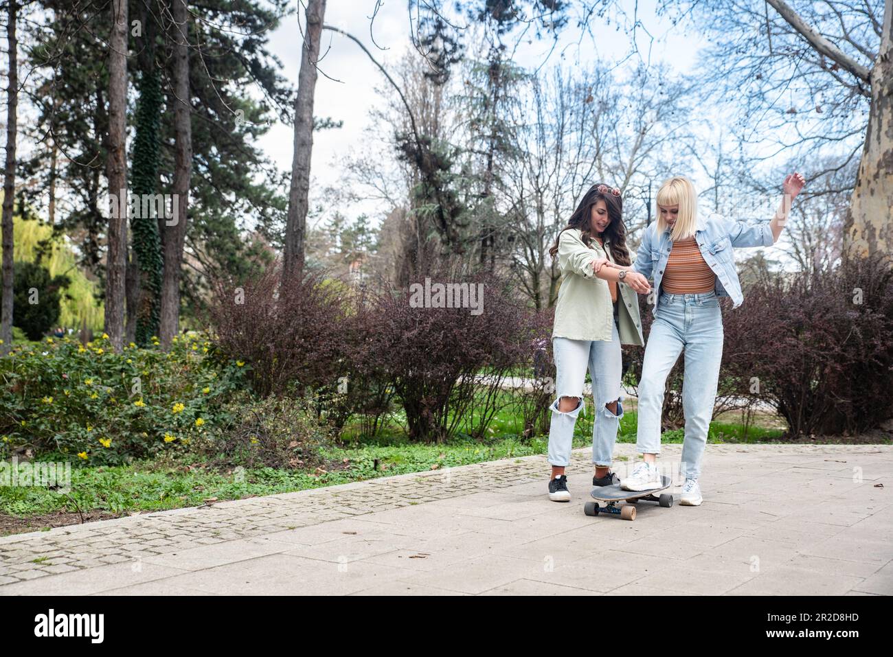 Two cool-looking hipster friends in park learning to ride a skateboard, having fun between studying or working, spending time together in a nice sunny Stock Photo