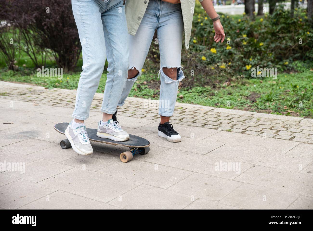 Two cool-looking hipster friends in park learning to ride a skateboard, having fun between studying or working, spending time together in a nice sunny Stock Photo