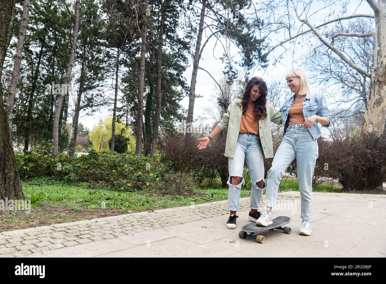 Two cool-looking hipster friends in park learning to ride a skateboard, having fun between studying or working, spending time together in a nice sunny Stock Photo