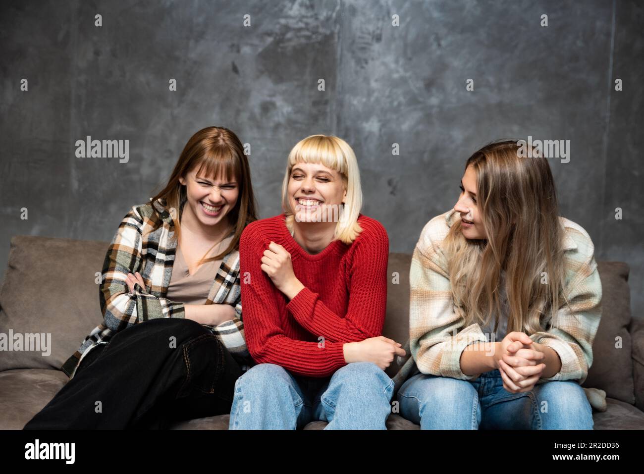 Three young female friends, roommates from college are sitting on the sofa and laughing because they are happy and successful in their lives. Stock Photo