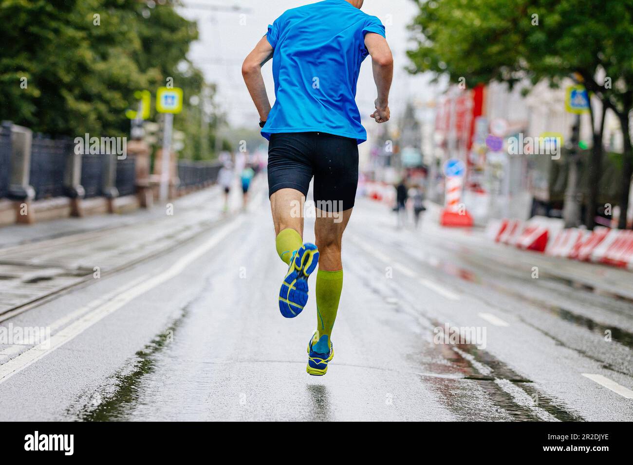 rear view male runner running city marathon on wet road, compression socks on feet, summer sports race Stock Photo