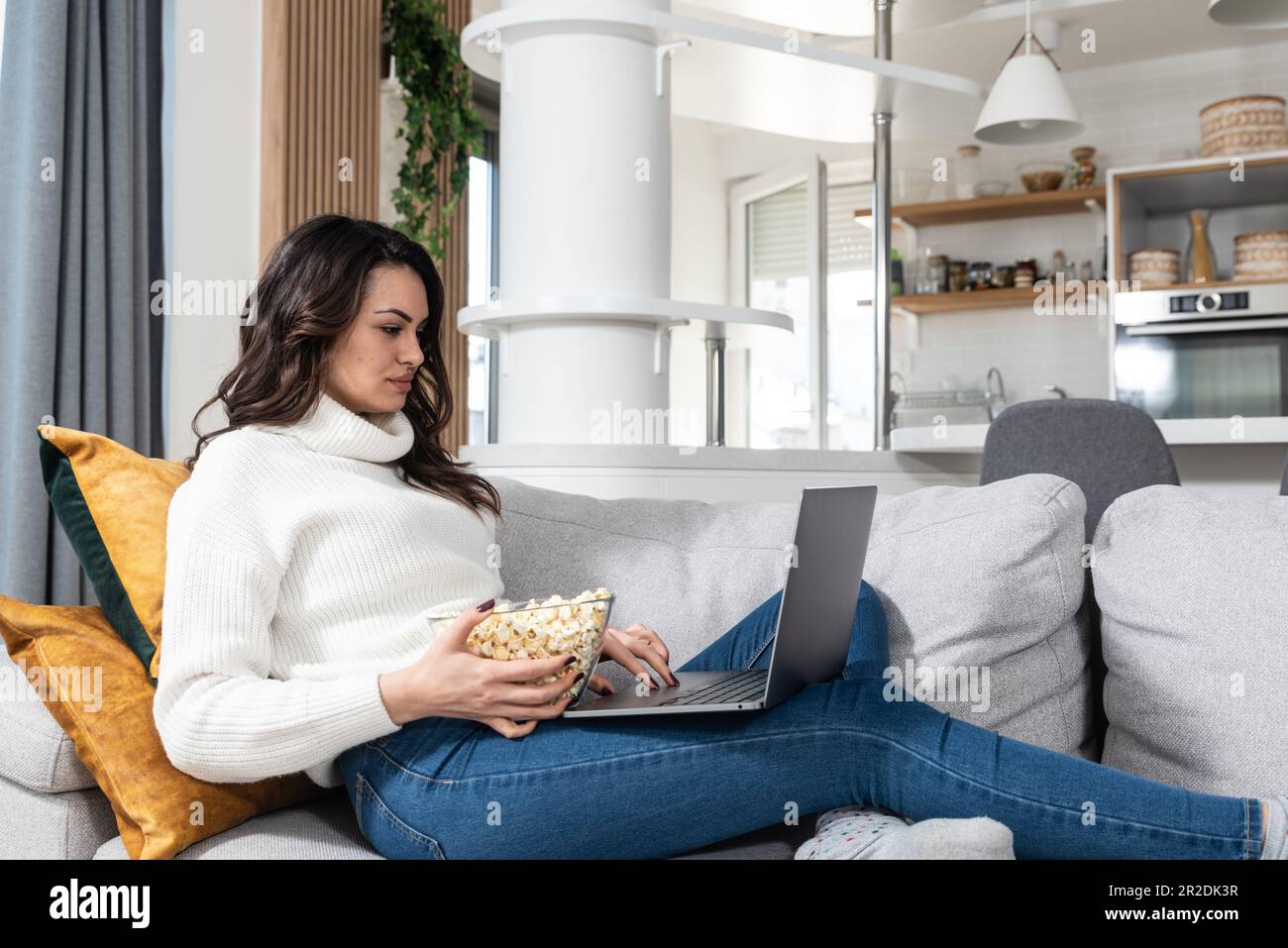 Happy young woman eating popcorn while watching her favorite series online on laptop computer in the evening at home on free day from work. Weekend le Stock Photo
