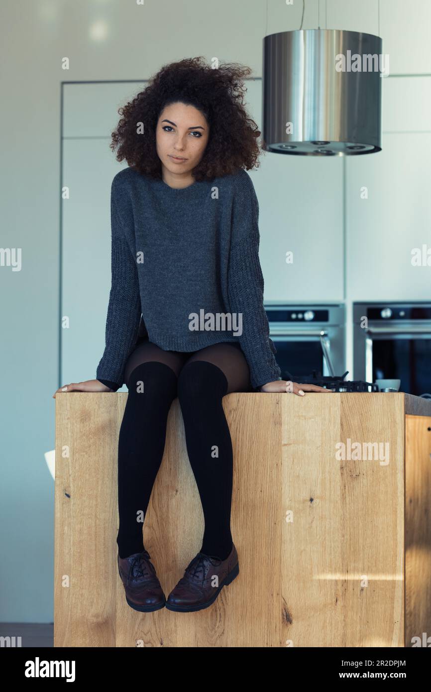 Curly-haired young woman in short skirt and warm sweater sits on her ultramodern kitchen island. The concept: love for cooking and cool lifestyle Stock Photo