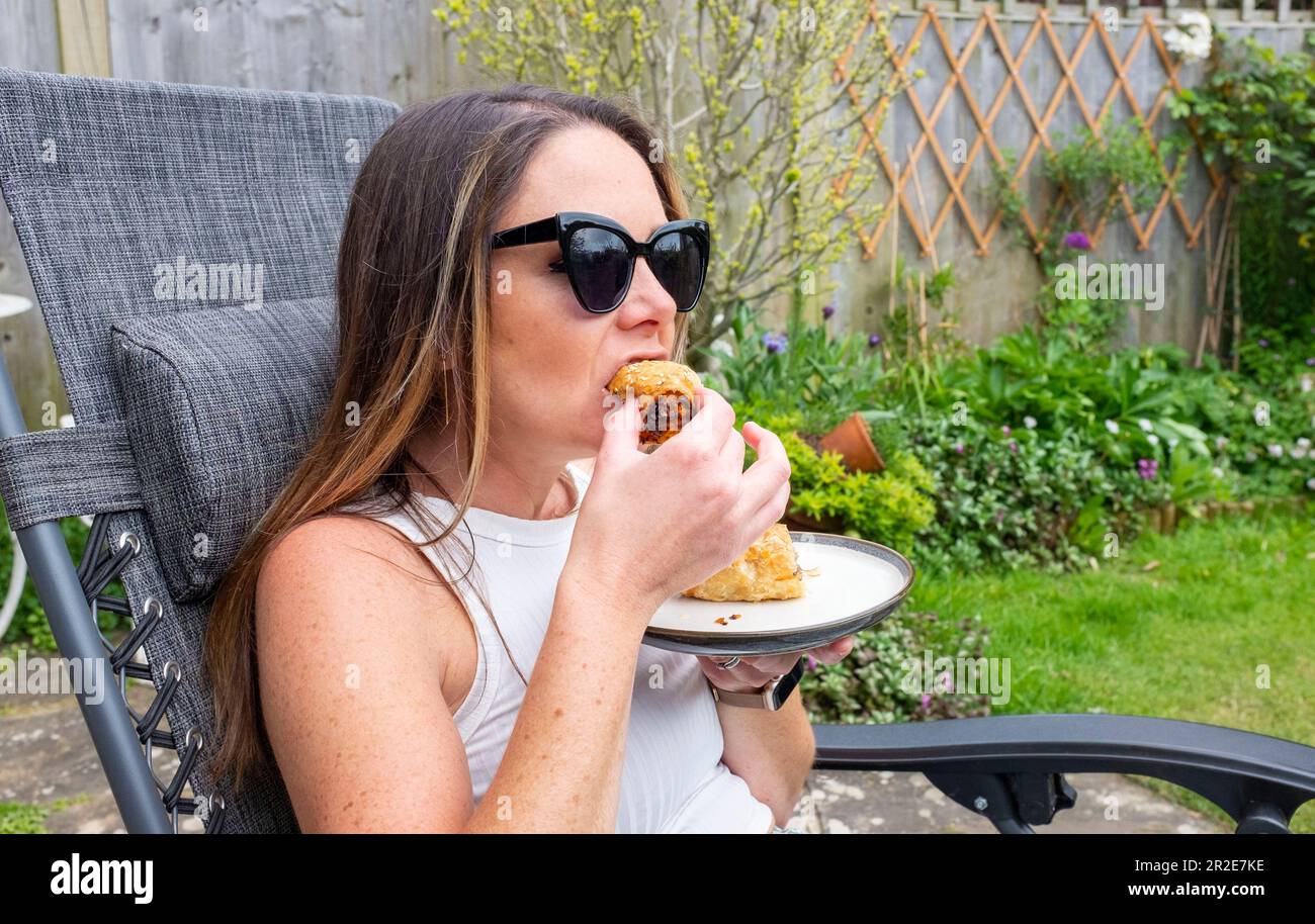 Young woman in her 30s eating a takeaway sausage roll in small urban garden in Spring sunshine  Credit Simon Dack Stock Photo