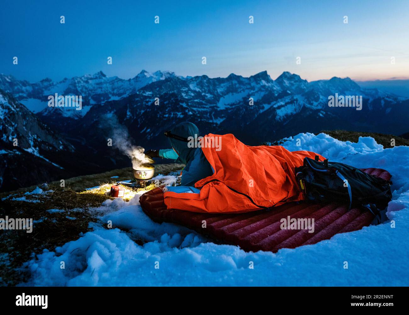Young woman lies in her bivouac sack on a sleeping pad in the snow and boils water with a gas