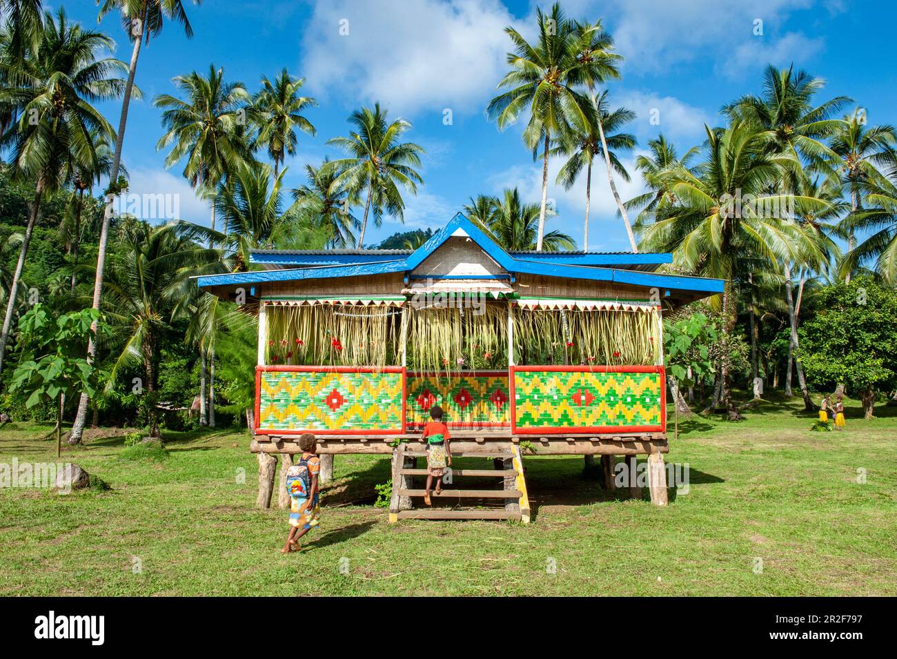 Two people enter a Selzen house with bright colors and patterns in a grassy landscape surrounded by palm trees, Garove Island, Vitu Islands, West New Stock Photo