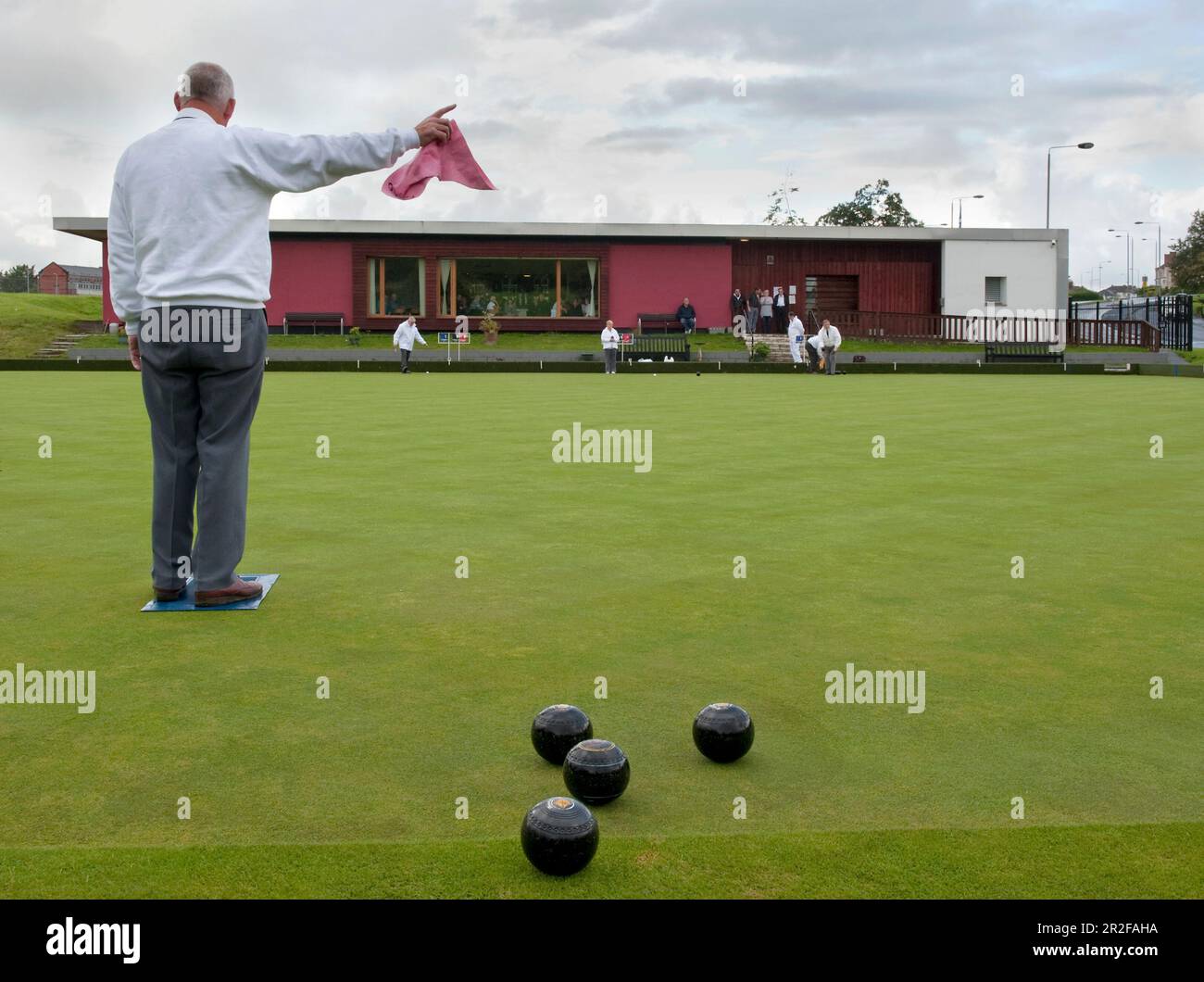 An older man waves a flag during bowls play in front of the red pavilion clubhouse at the Balornock lawn bowling green in Glasgow, Scotland, UK Stock Photo
