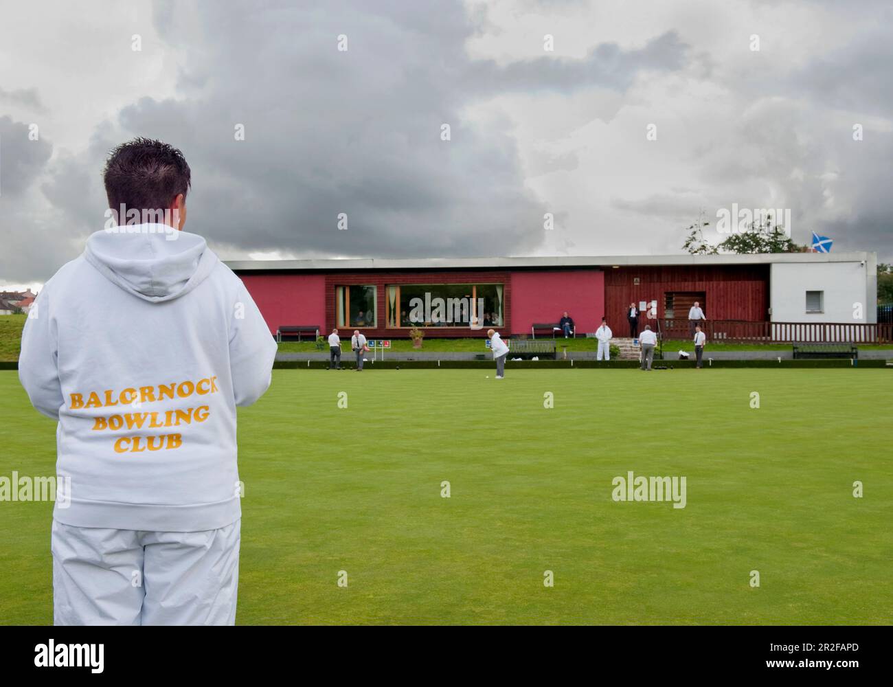 A woman wearing a white club hoodie plays bowls in front of the red pavilion clubhouse at the Balornock lawn bowling green in Glasgow, Scotland, UK Stock Photo