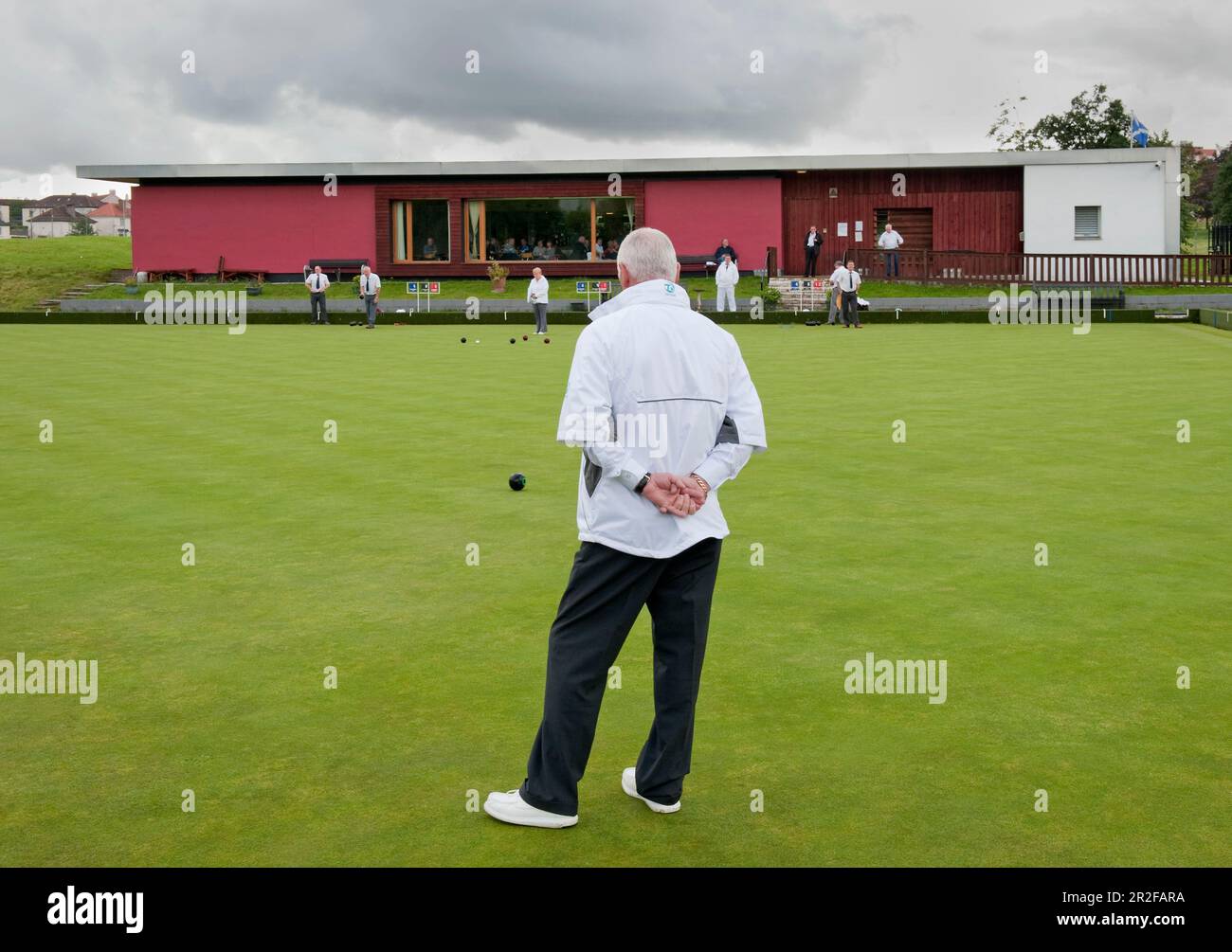 A man stands in the foreground of bowls in play in front of the red pavilion clubhouse at the Balornock lawn bowling green in Glasgow, Scotland, UK Stock Photo