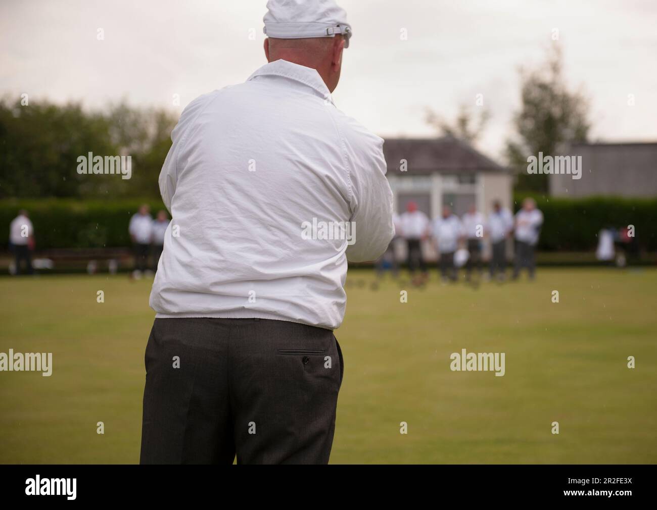 A bald man wearing a hat play bowls in front of a crowd outside the pavilion at the Mauchline lawn bowling green in Mauchline; Ayrshire, Scotland, UK Stock Photo