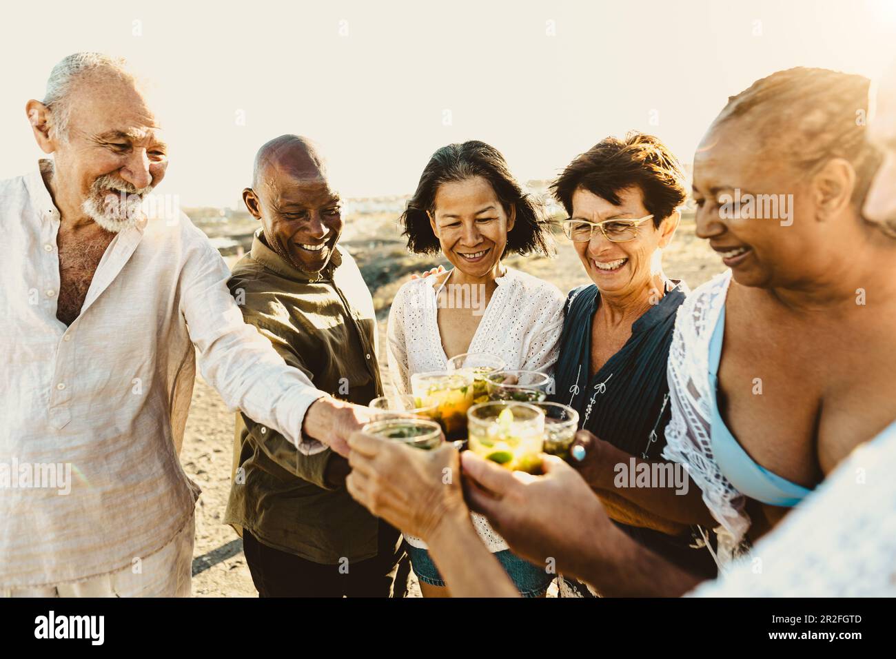 Happy multiracial senior friends having fun drinking and toasting mojitos on the beach during sunset time - Elderly people enjoying summer holidays Stock Photo