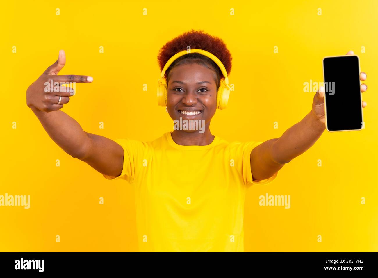 Young african american woman isolated on a yellow background with mobile pointing gesture, studio shoot Stock Photo