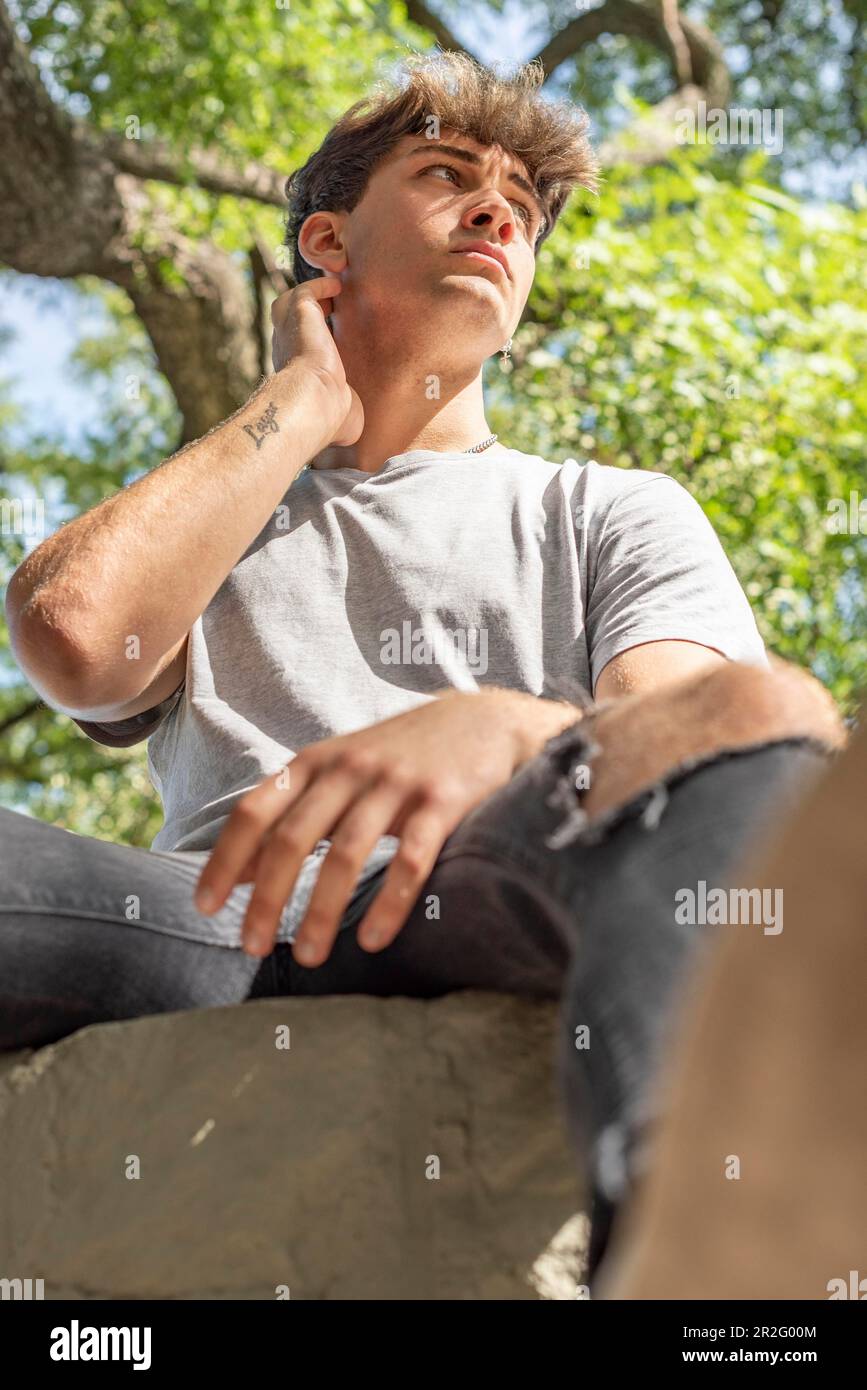 Low angle view of a young man, serious, sitting on a tree while looking away Stock Photo