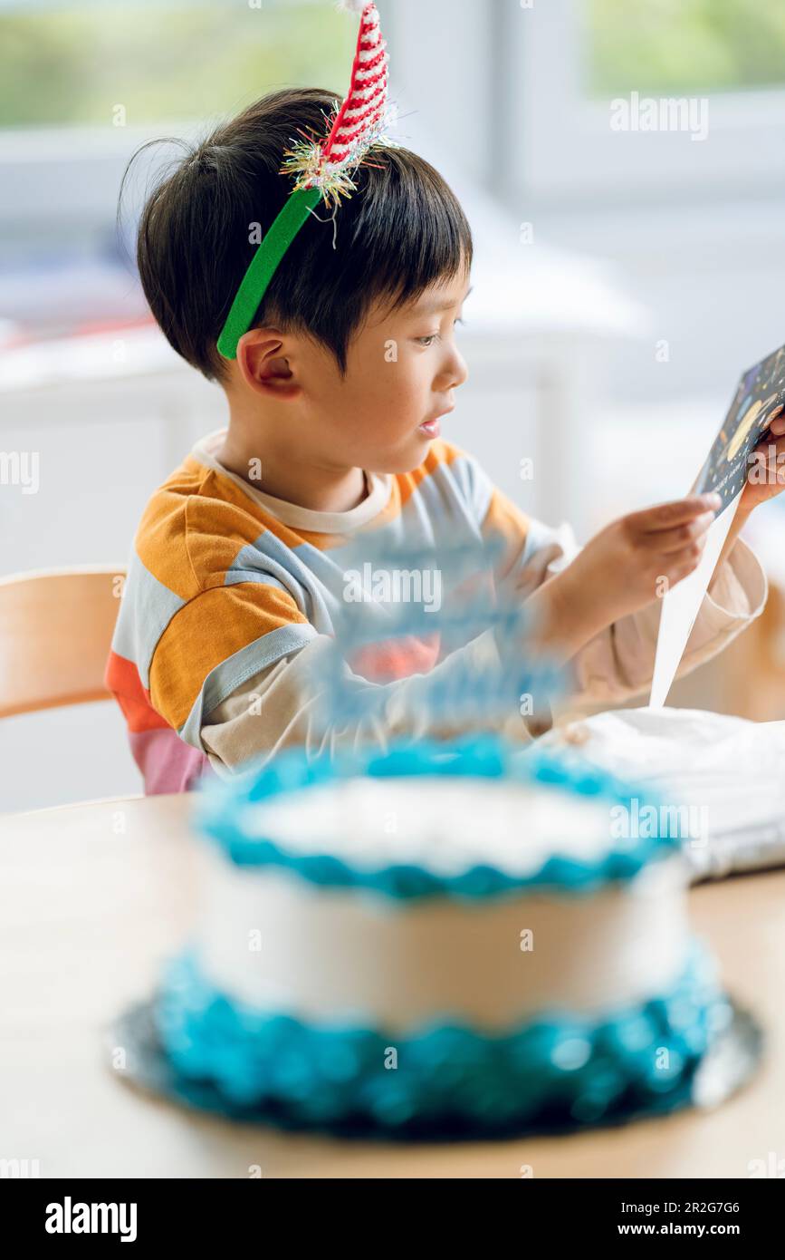 A boy celebrates his birthday. He reads his birthday card with a smile. a cake with blue creamy decoration on the table. Stock Photo