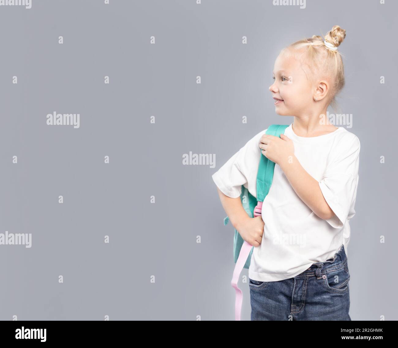 Elementary school girl with blond hair and messy buns, holding backpack and dressed in white t-shirt looking sideways on grey background Stock Photo