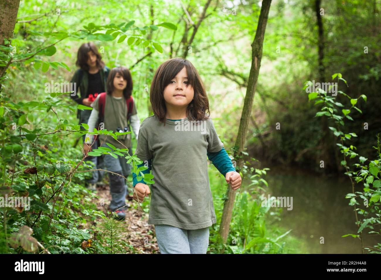 Three siblings walking and exploring the forest alongside a stream. In the foreground, the youngest sibling is leading the way. Stock Photo
