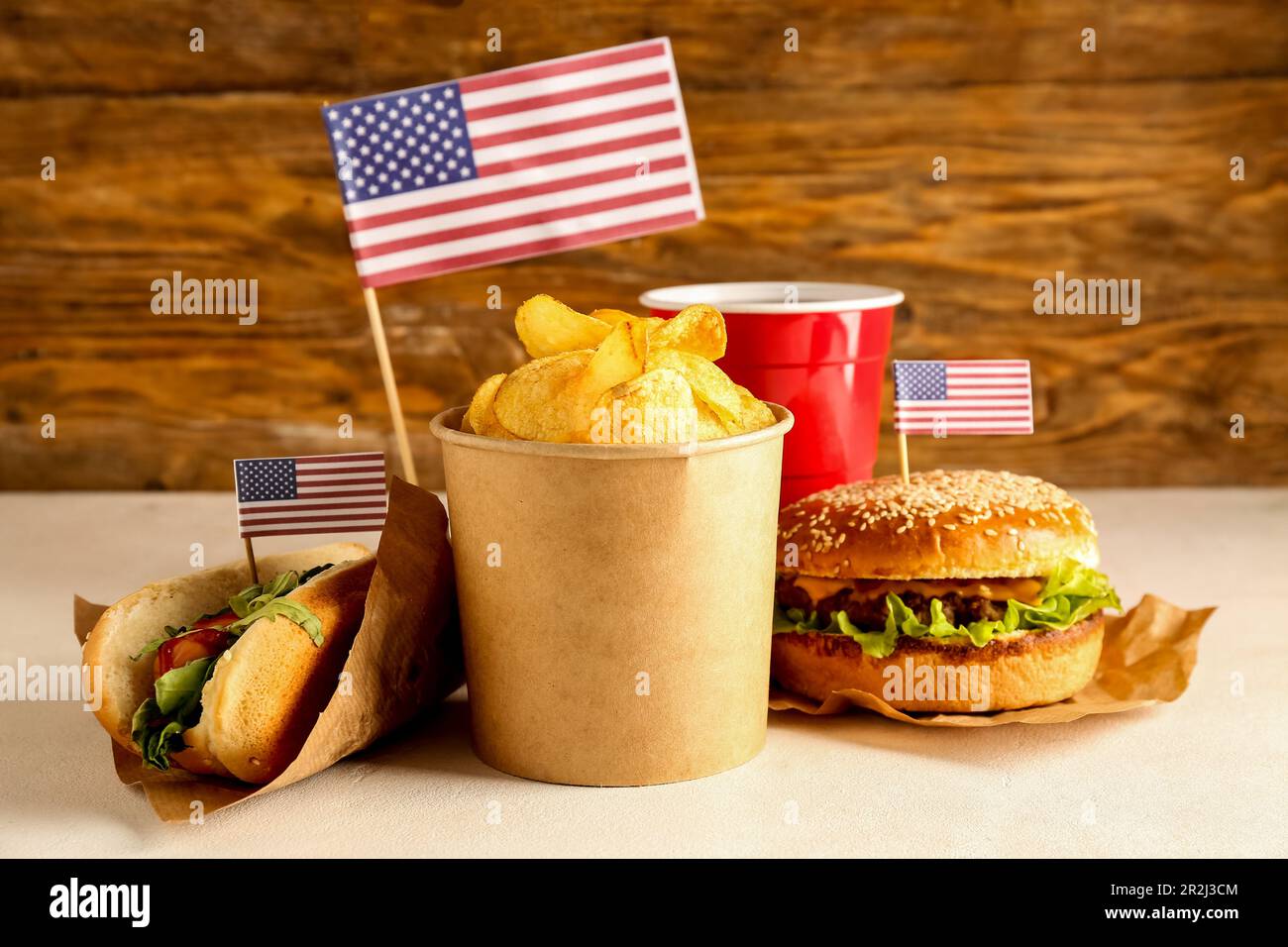 Tasty burger, potato chips, hot dog and cup of drink on light table against wooden background. Memorial Day celebration Stock Photo