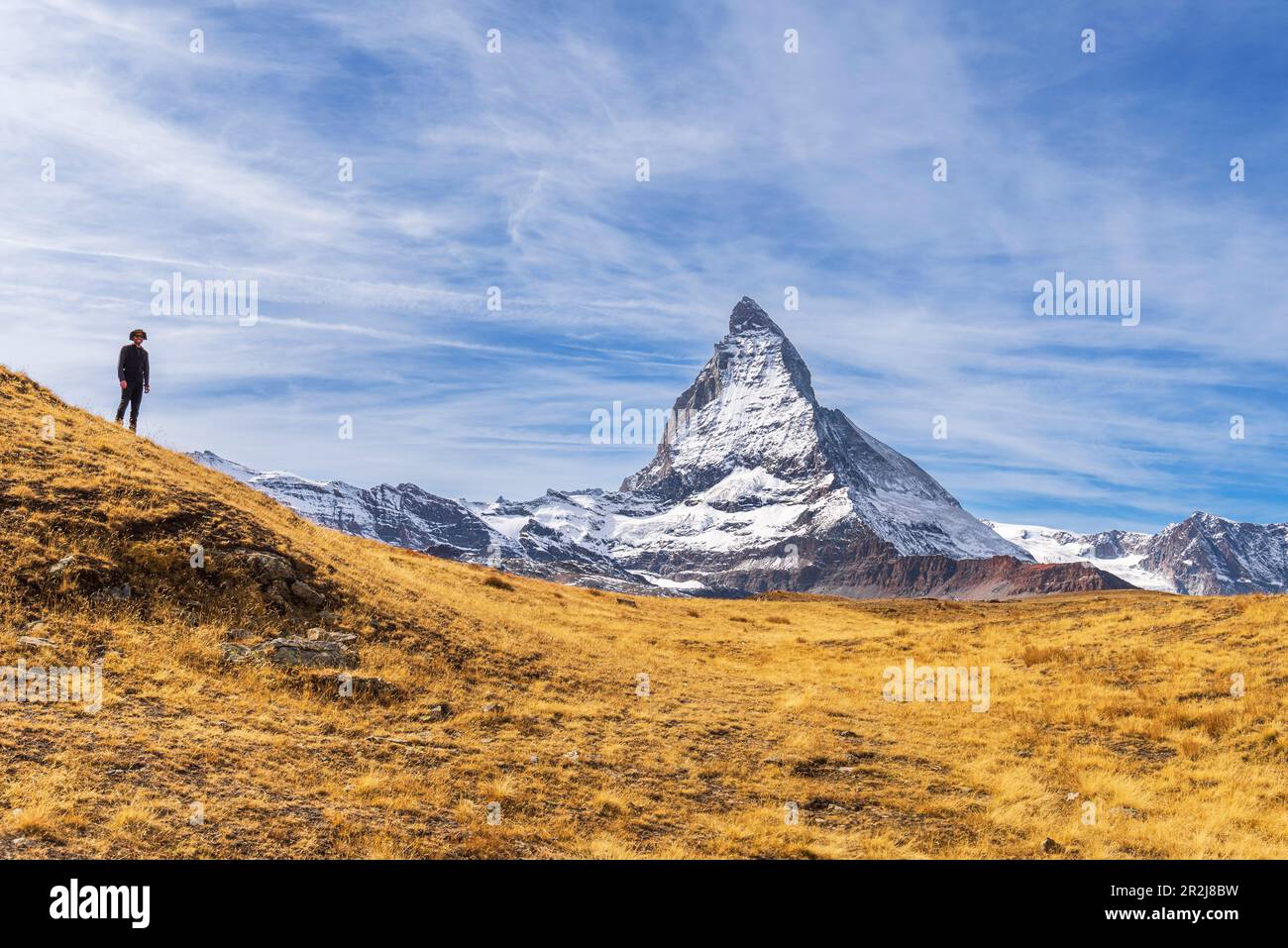 Hiker stands in front of the iconic shape of Matterhorn among yellow grasses, Riffelalp, Zermatt, Valais Canton, Swiss Alps, Switzerland, Europe Stock Photo