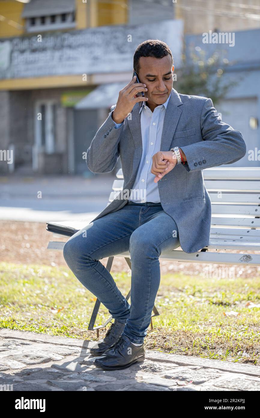 Young latin man sitting on a square bench talking on mobile phone and looking at his watch. Stock Photo