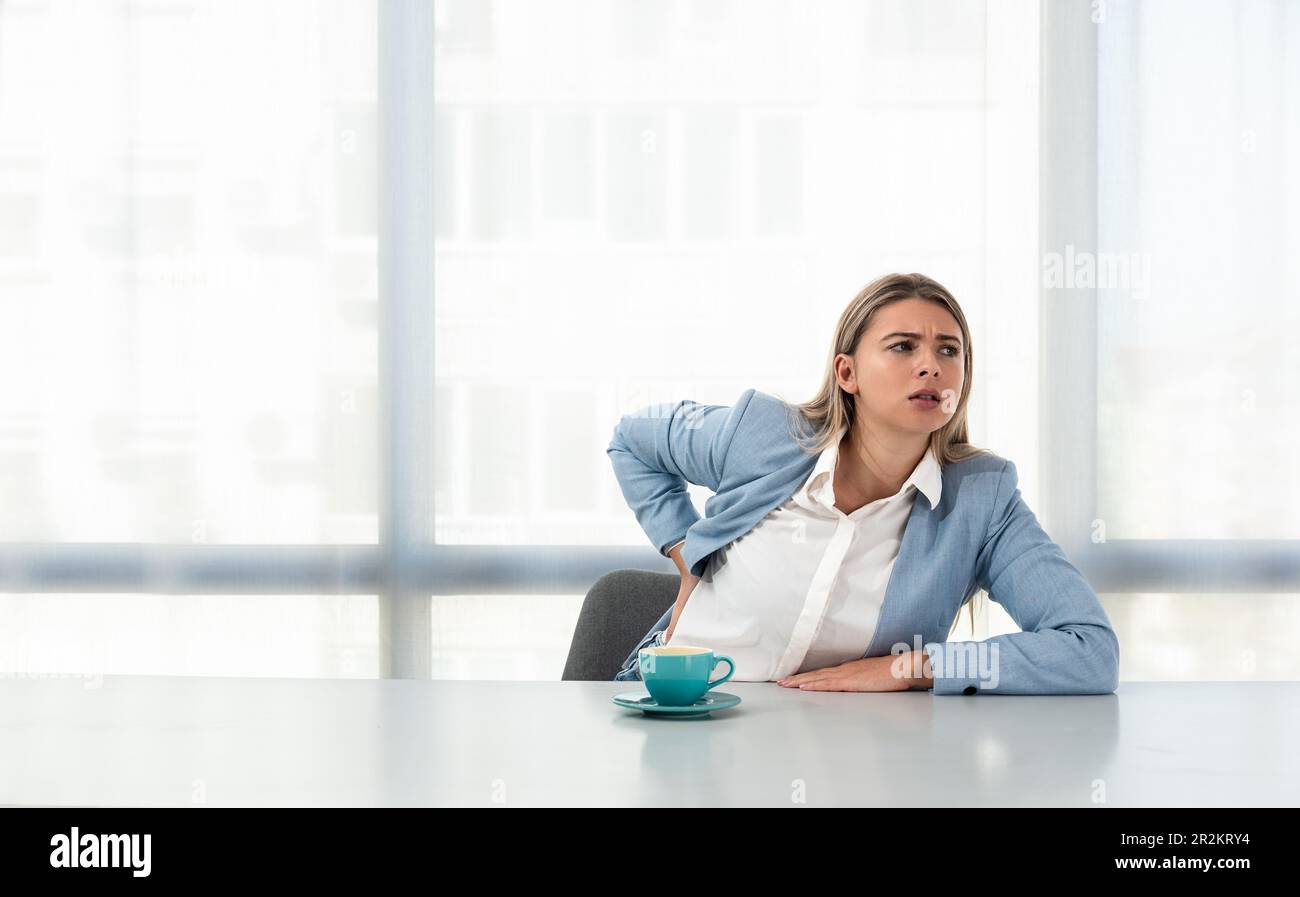Young business woman having back pain ache from bad posture while sitting at desk in office on non ergonomic chair at the work place in formal wear wi Stock Photo