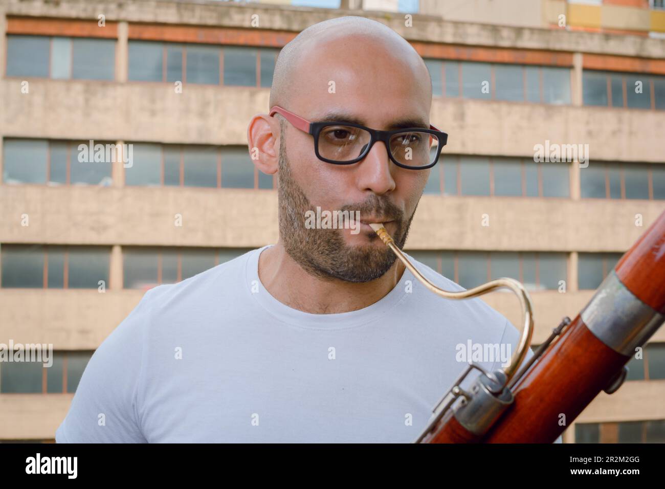 closeup portrait of young latino man with glasses, beard and bald, wearing white t-shirt, standing playing bassoon on apartment balcony with urban bui Stock Photo
