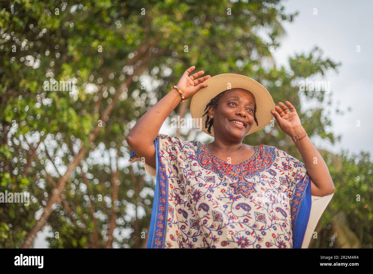 Happy African woman holding her summer hat and smiling while relaxing outdoors Stock Photo