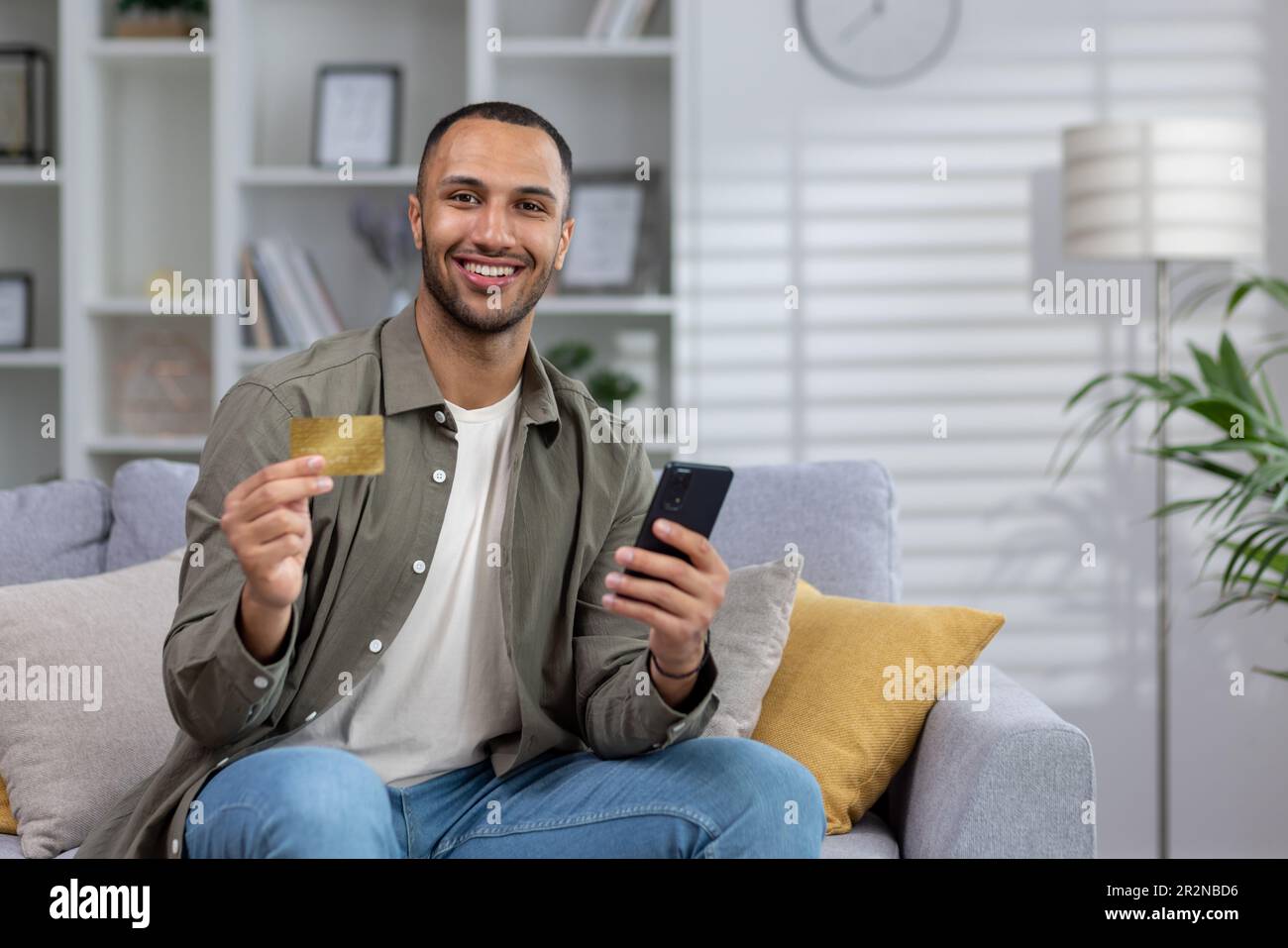 Online shopping. Portrait of a young African American man is sitting on the couch at home, holding a credit card and a phone. Smiling at the camera. Stock Photo