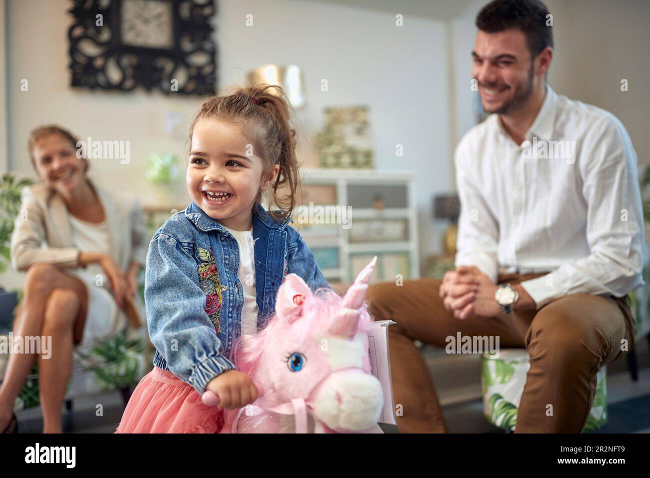 Cute toddler girl riding a pink unicorn rocking horse in a furniture shop, lovely family in a home decor shop together. Shopping, lifestyle, family co Stock Photo