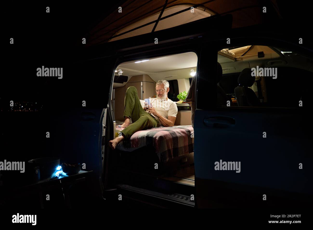 Old man traveler sitting in camper van using mobile in camping at night
