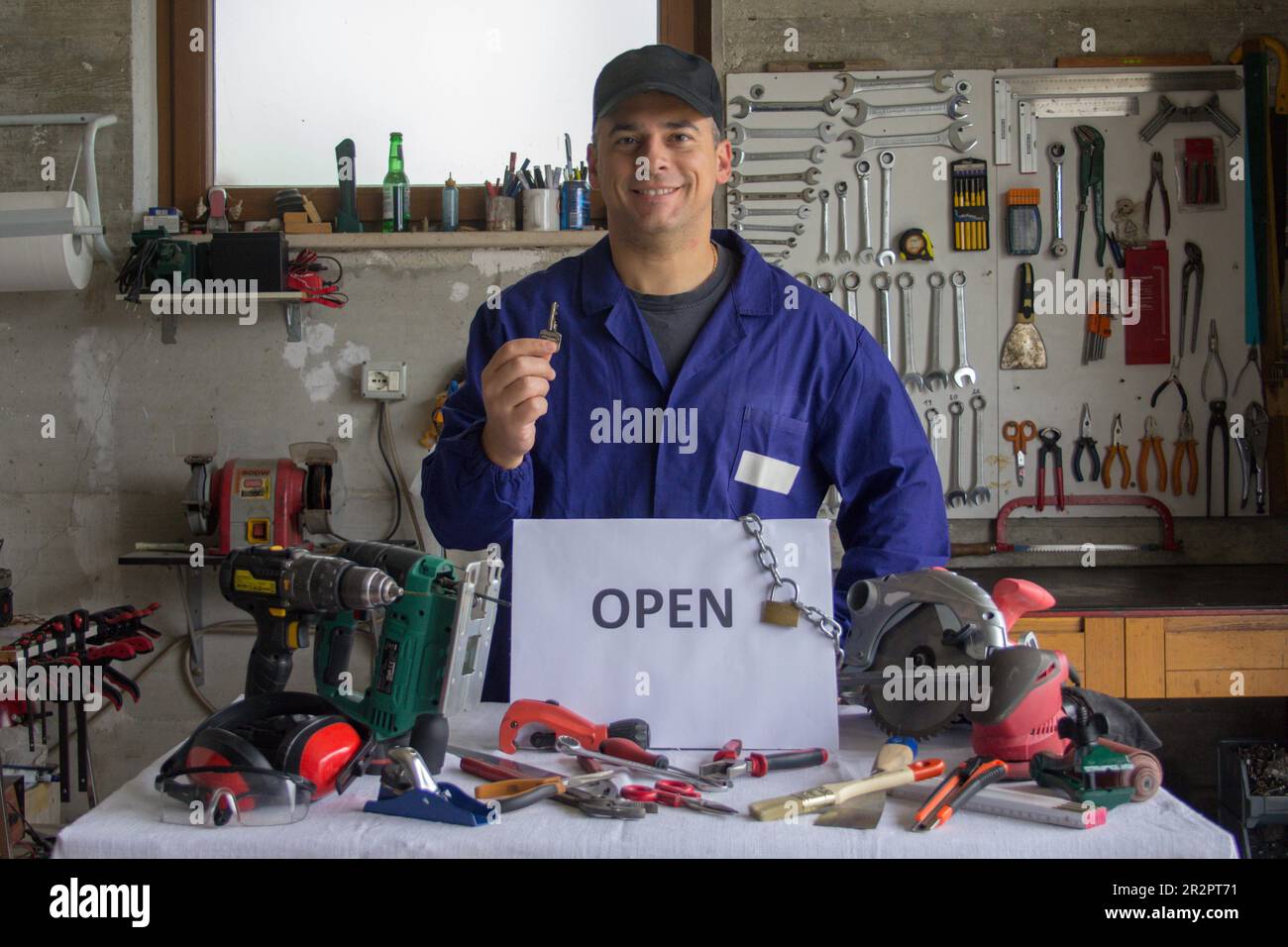Image of a smiling handyman in his workshop with an OPEN sign and various work tools. Reopening of work activities and work development Stock Photo