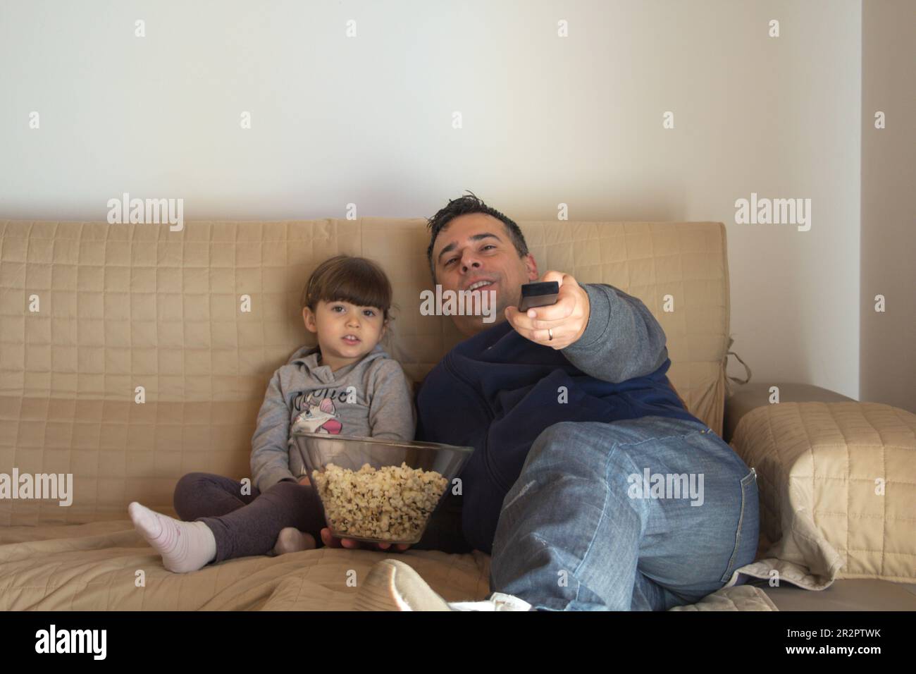 Father and daughter lying on the sofa at home eating popcorn and watching TV. Dad who zaps with the TV remote control. Stock Photo
