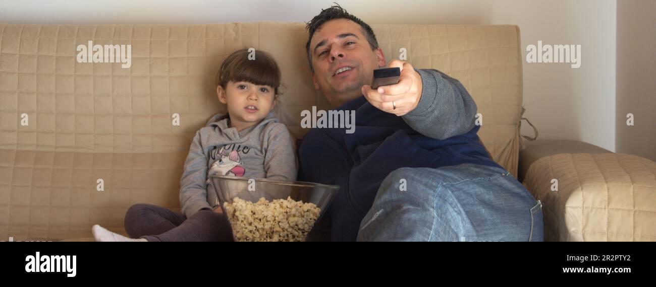 Father and daughter lying on the sofa at home eating popcorn and watching TV. Dad who zaps with the TV remote control. Horizontal banner Stock Photo